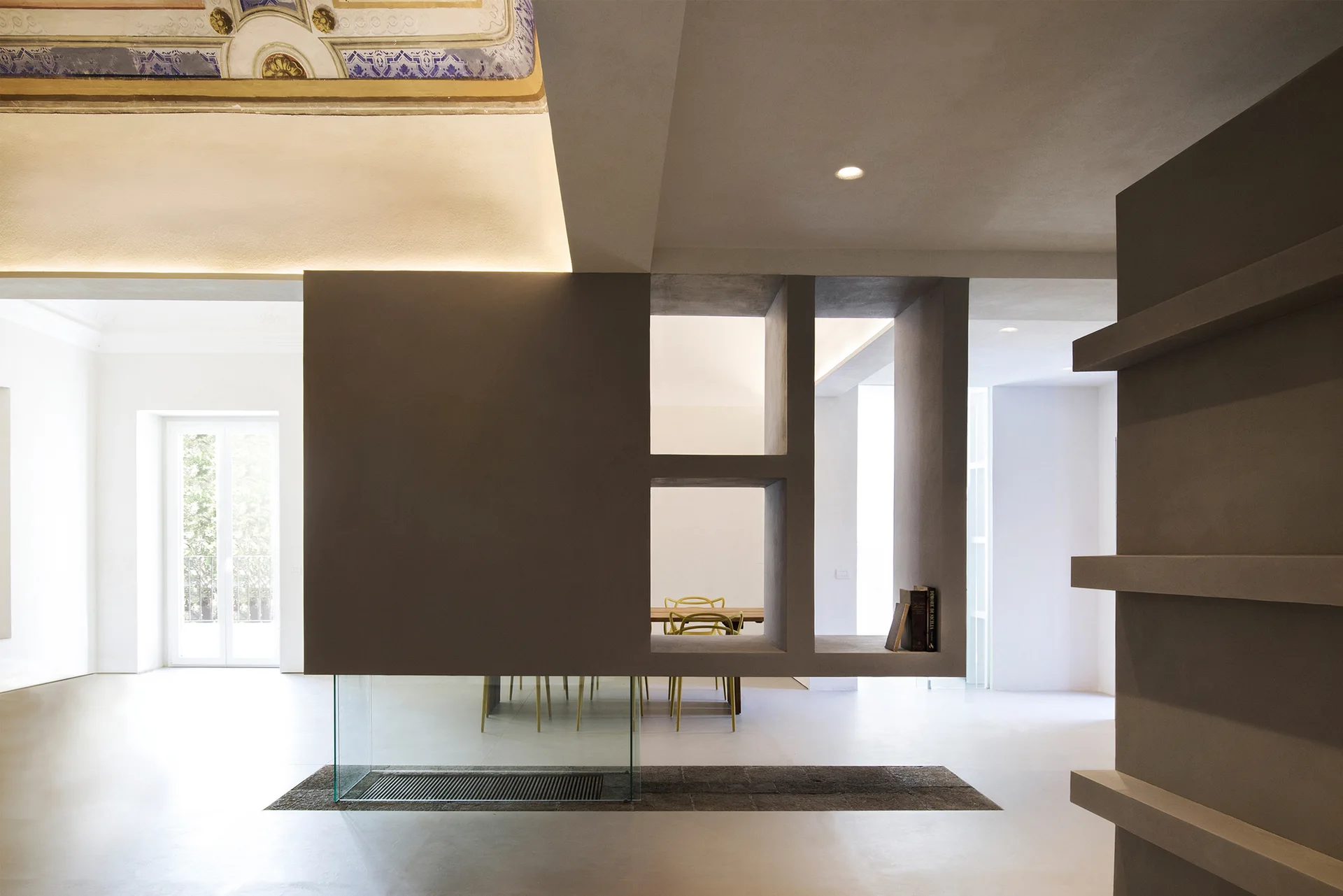 A dark sculptural room divider with cutouts stands on a glass base, separating an off-white hallway from a dining room with yellow chairs.
