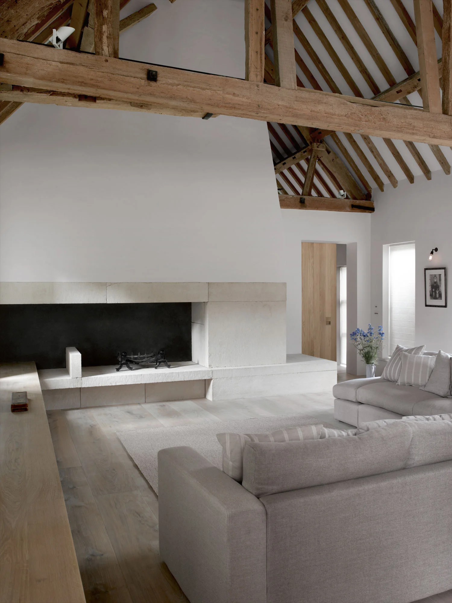 A minimalist living room with grey sofas arranged on a light rug before a massive stone fireplace under a vaulted ceiling with rustic wood beams.