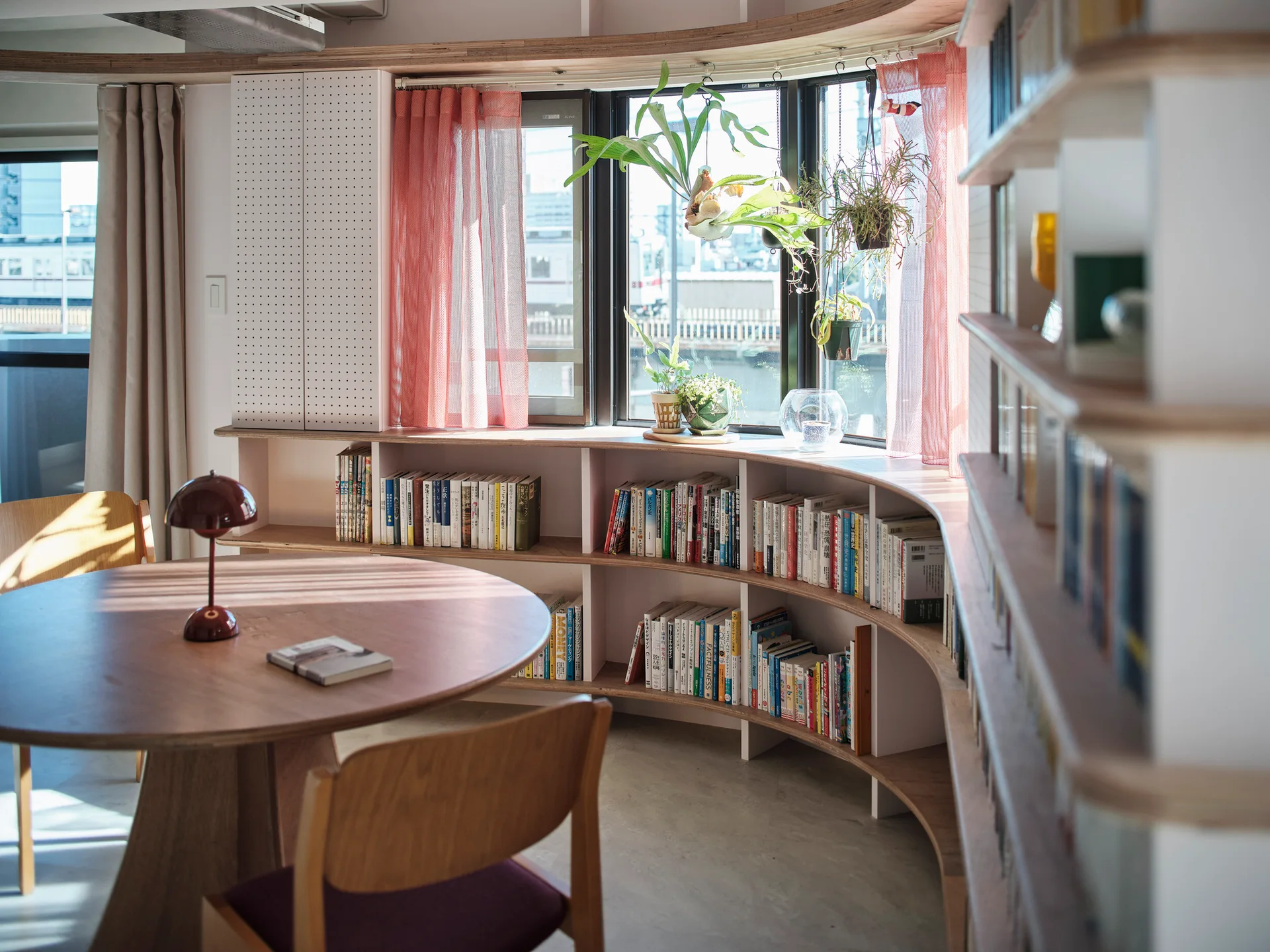 A curved plywood bookshelf filled with books wraps around a window wall near a round wooden table with a small red lamp.