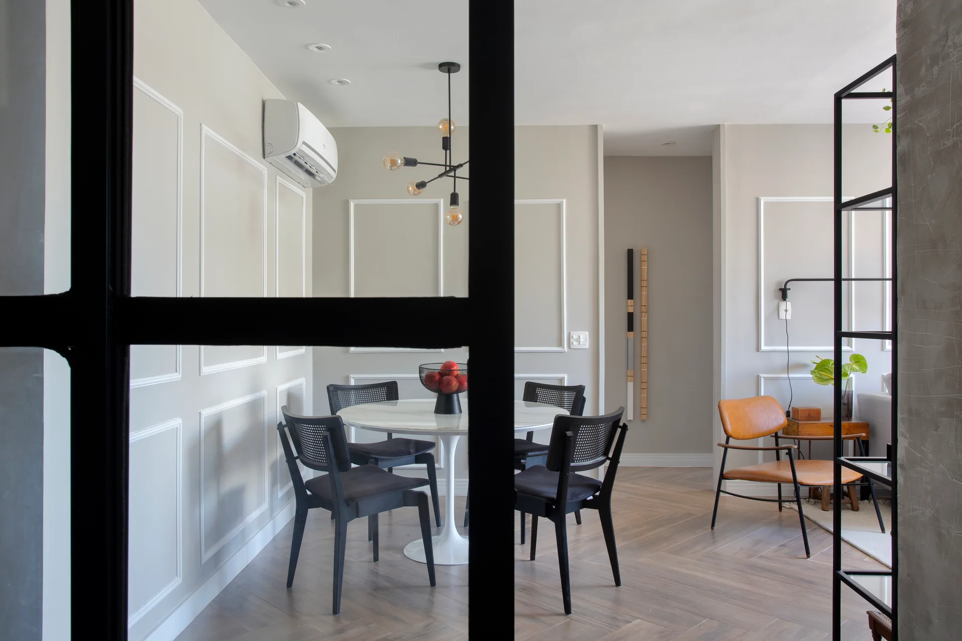 A dining room seen through a black metal partition features a white pedestal table, black cane-back chairs, gray paneled walls, and herringbone floors.