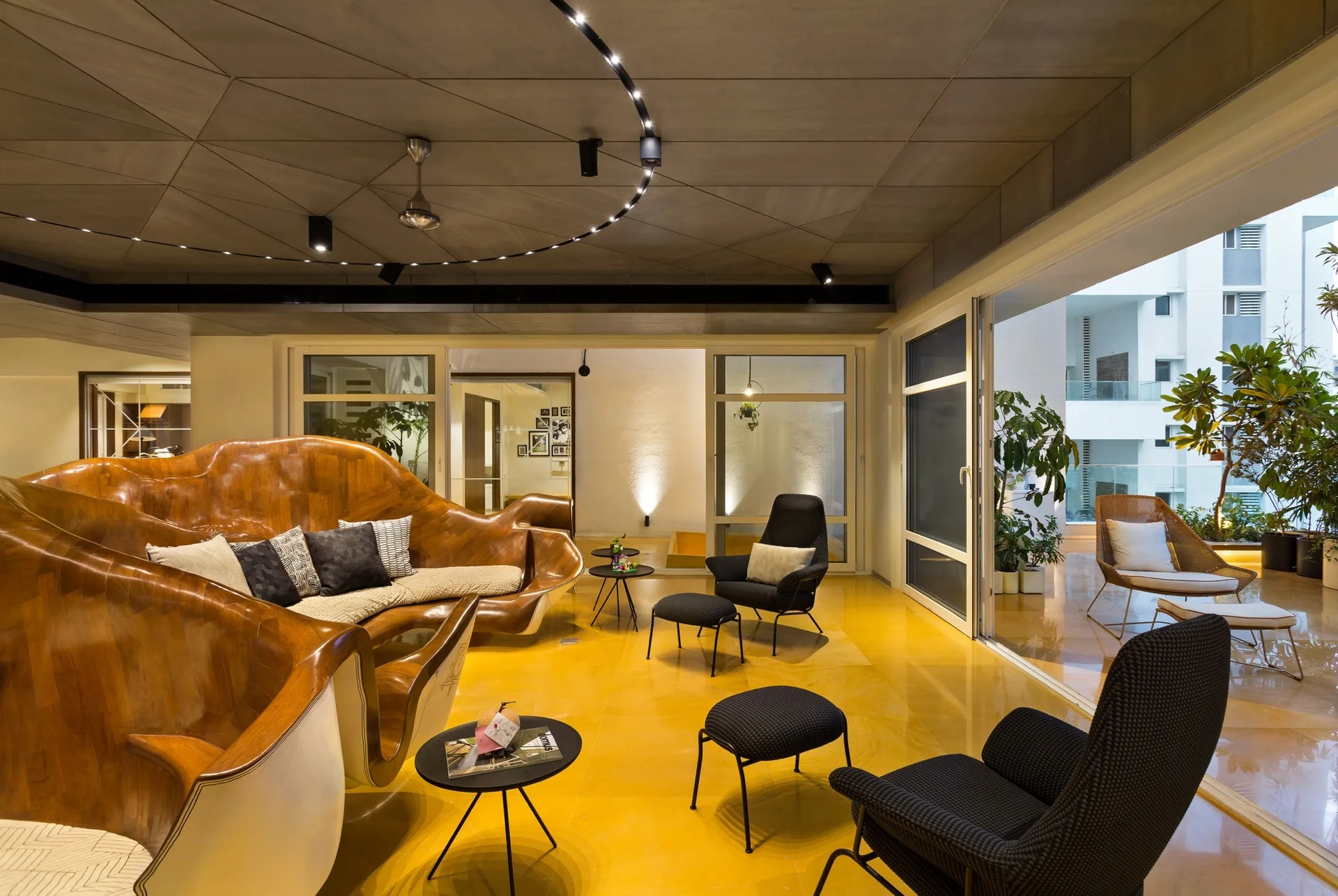 A modern living space featuring a sculptural wooden sofa, black armchairs, circular track lighting, and reflective yellow flooring.