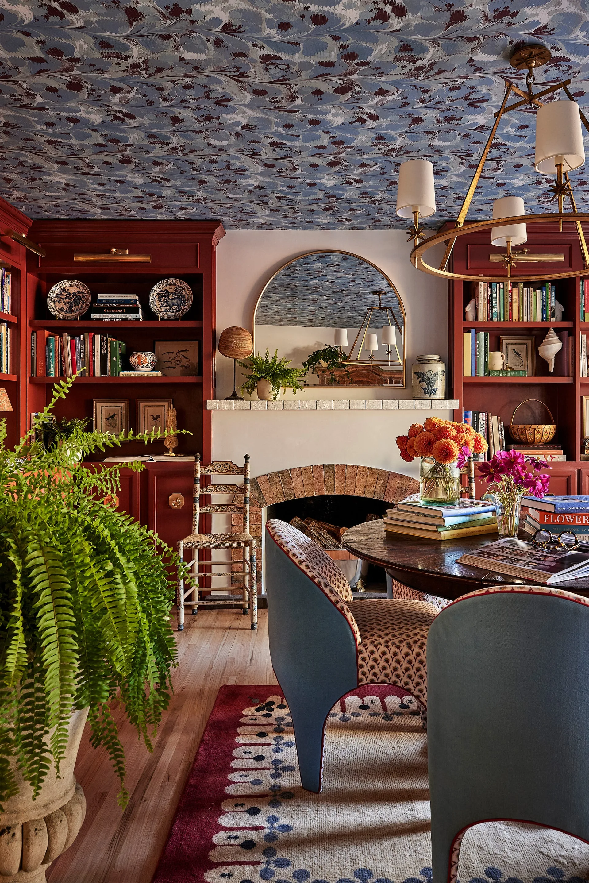 A cozy library features deep red built-in bookshelves flanking a brick fireplace with an arched mirror, beneath a blue marbled wallpapered ceiling.