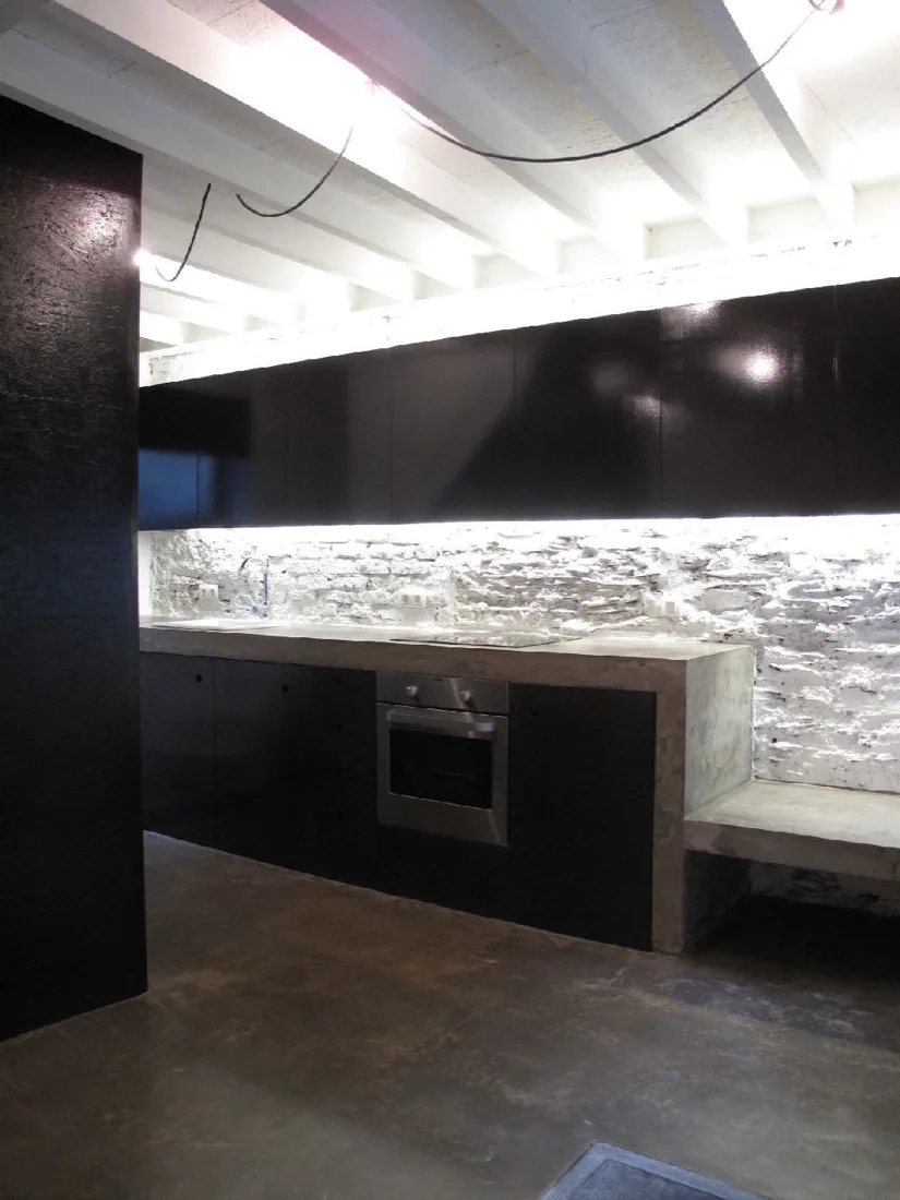 A modern industrial kitchen featuring black flat-panel cabinets, a cast concrete countertop, a textured white stone backsplash, and exposed ceiling beams.