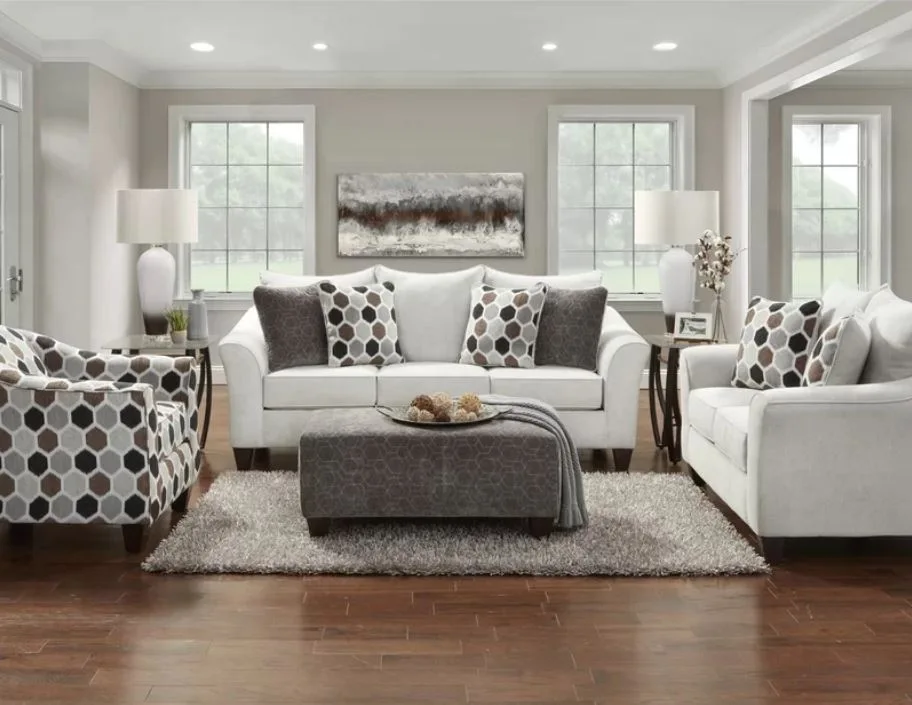 A symmetrically arranged living room features a white sofa and loveseat, a grey patterned ottoman, and a bold geometric armchair on a shag rug.