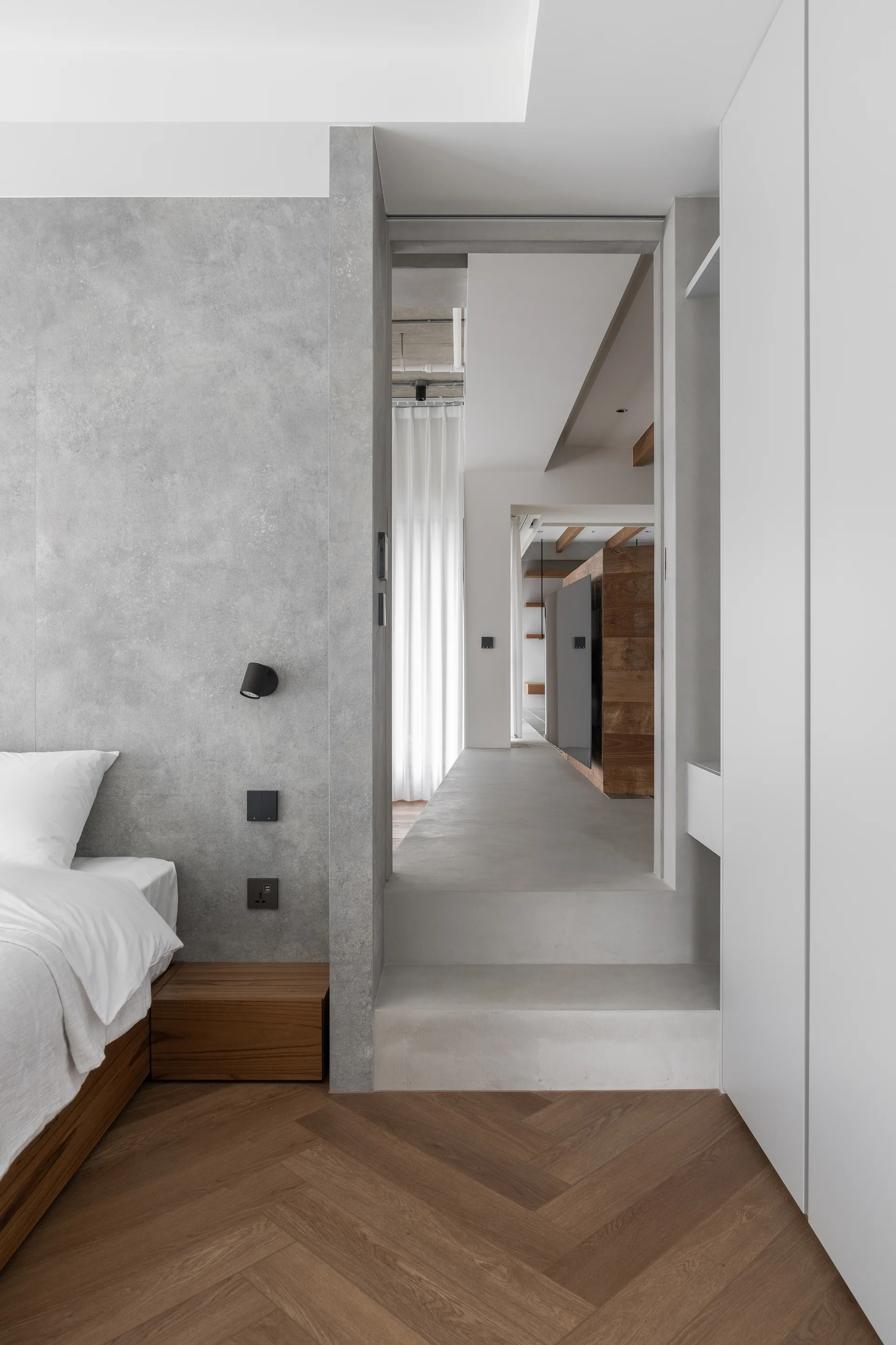 A minimalist bedroom features a grey textured wall, herringbone wood floors, a low bed, and a raised concrete step leading to a hallway.