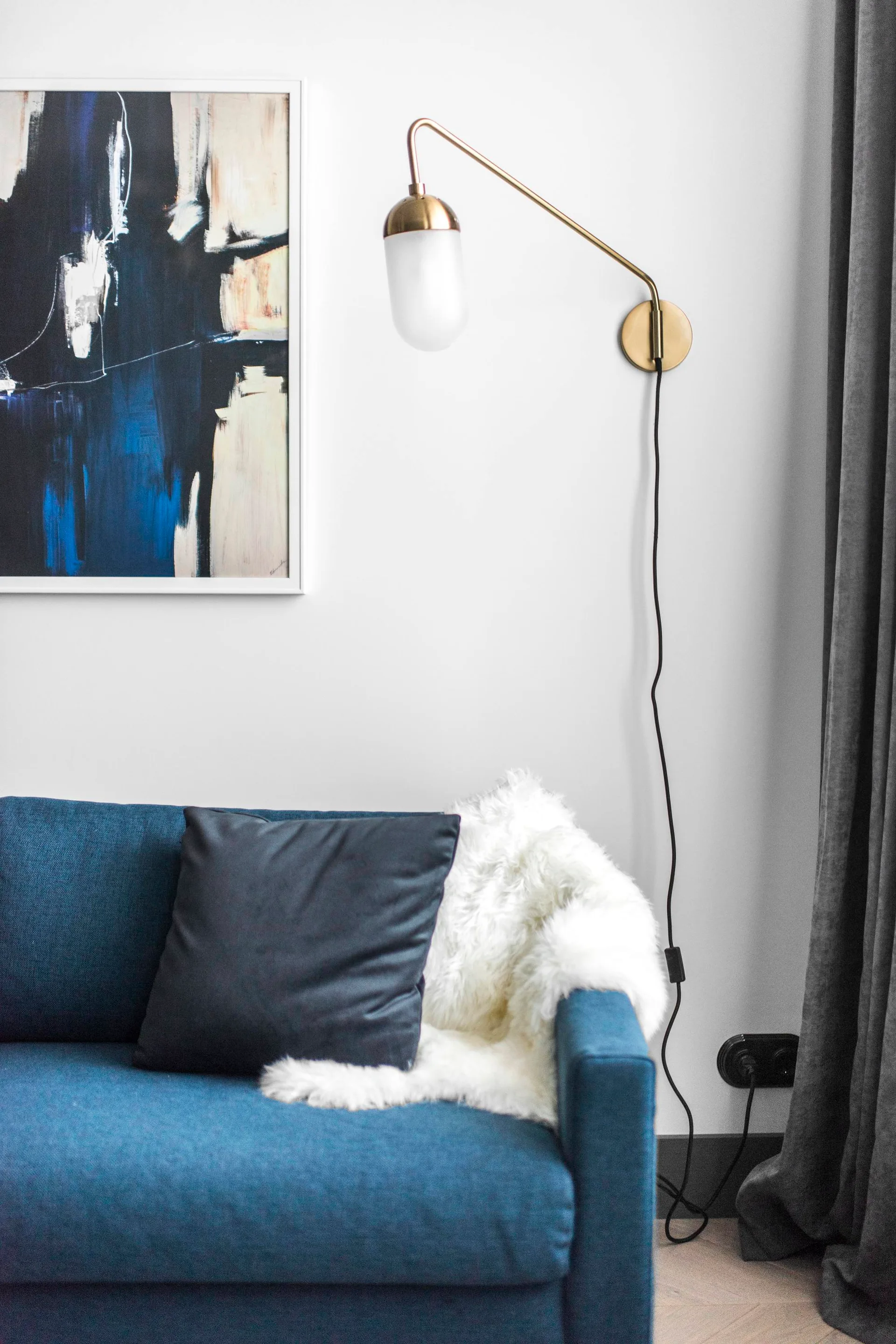 A blue fabric sofa styled with a white faux fur throw, a dark pillow, a brushed brass wall sconce, and framed abstract artwork.