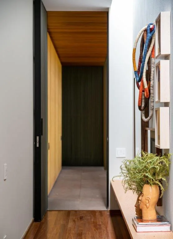 A narrow white hallway features a low wood console table with a face-shaped vase and colorful coiled fiber art hanging on the wall.