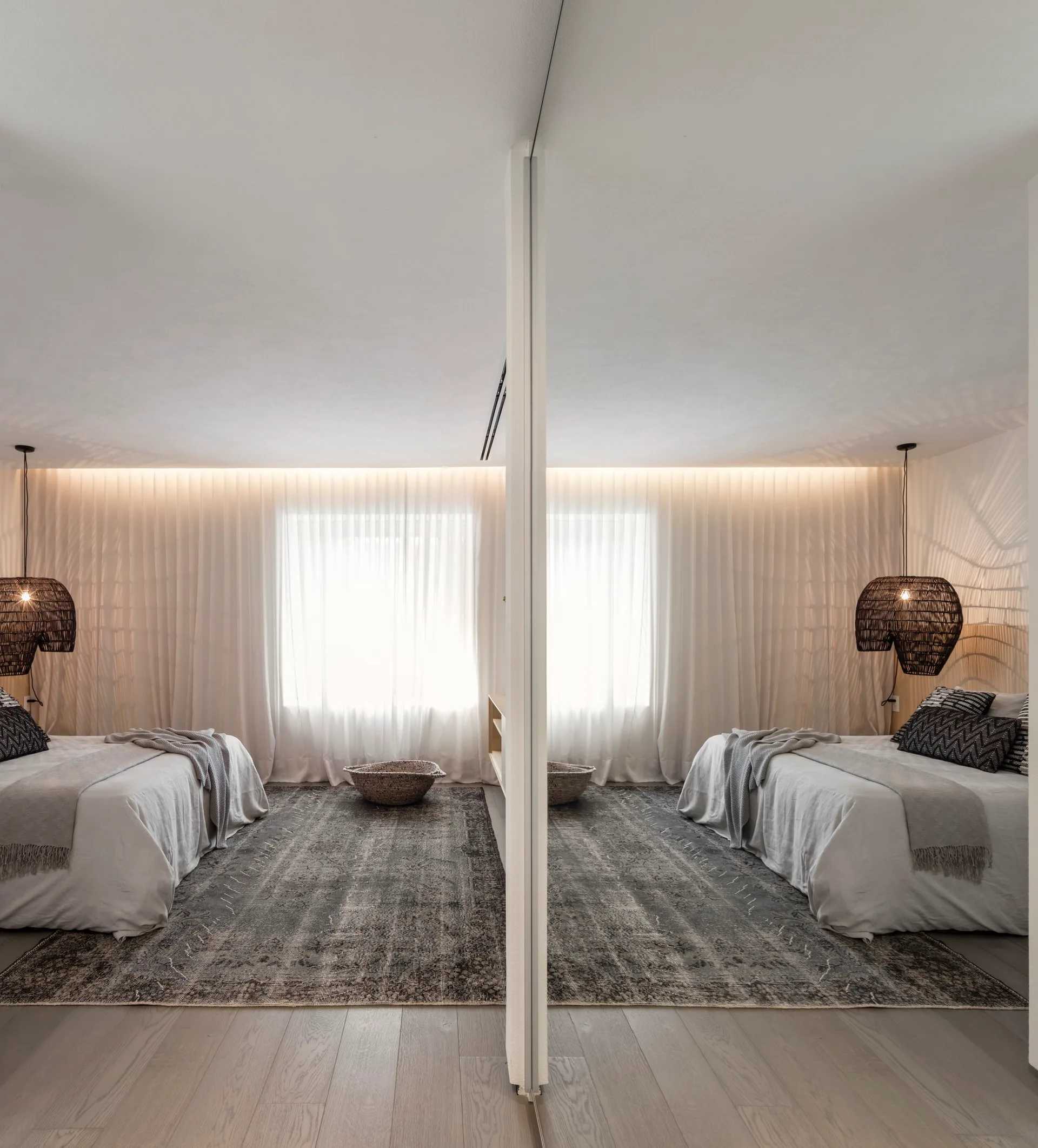 A contemporary bedroom featuring a central mirrored partition, two beds with grey linens, and sheer curtains lit by cove lighting.
