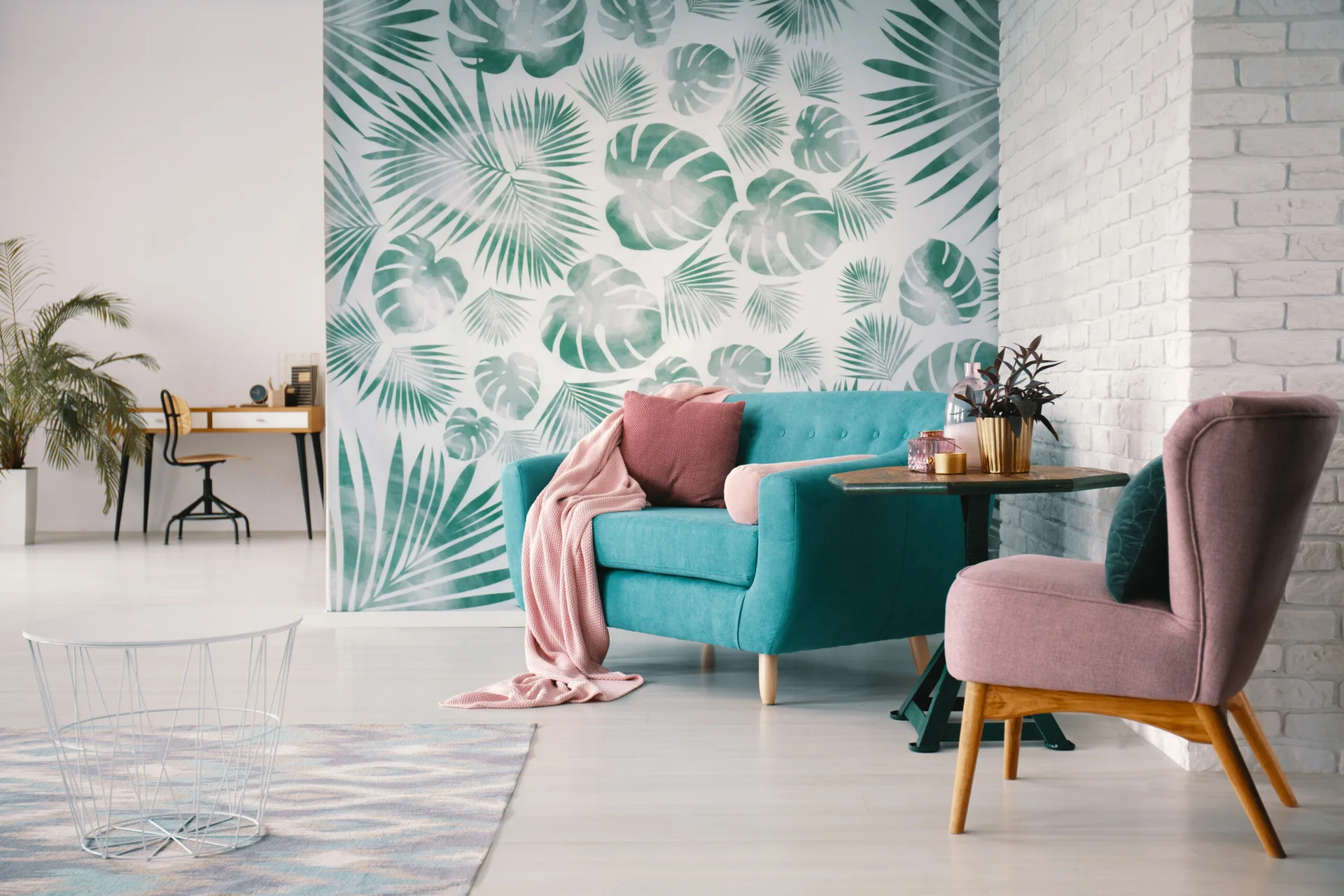 A teal sofa and pink armchair sit in a living room featuring a tropical green leaf wallpaper wall and a white painted brick wall.