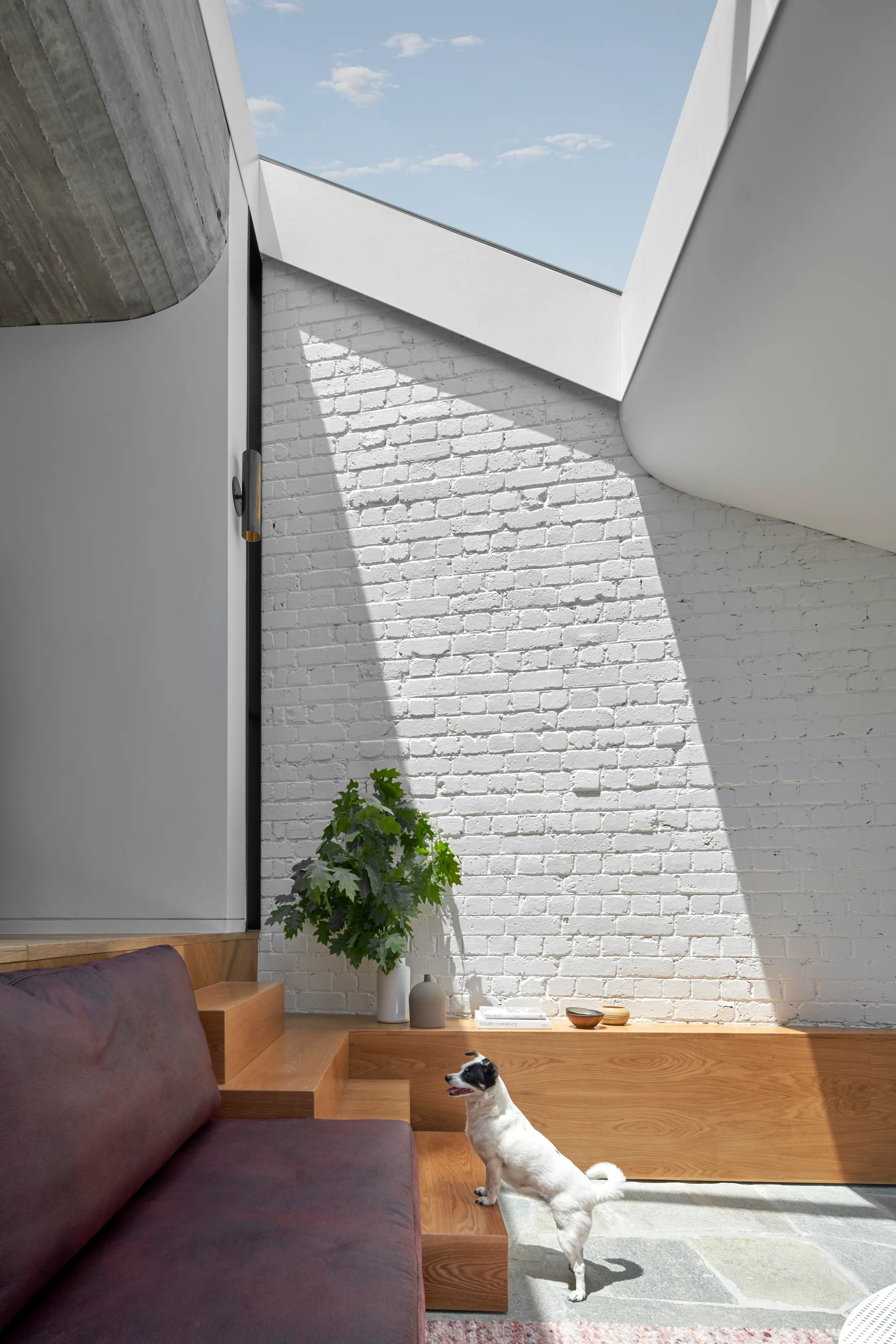 A sunlit living area features a painted white brick wall beneath a large skylight, a custom wooden bench, and flagstone flooring.