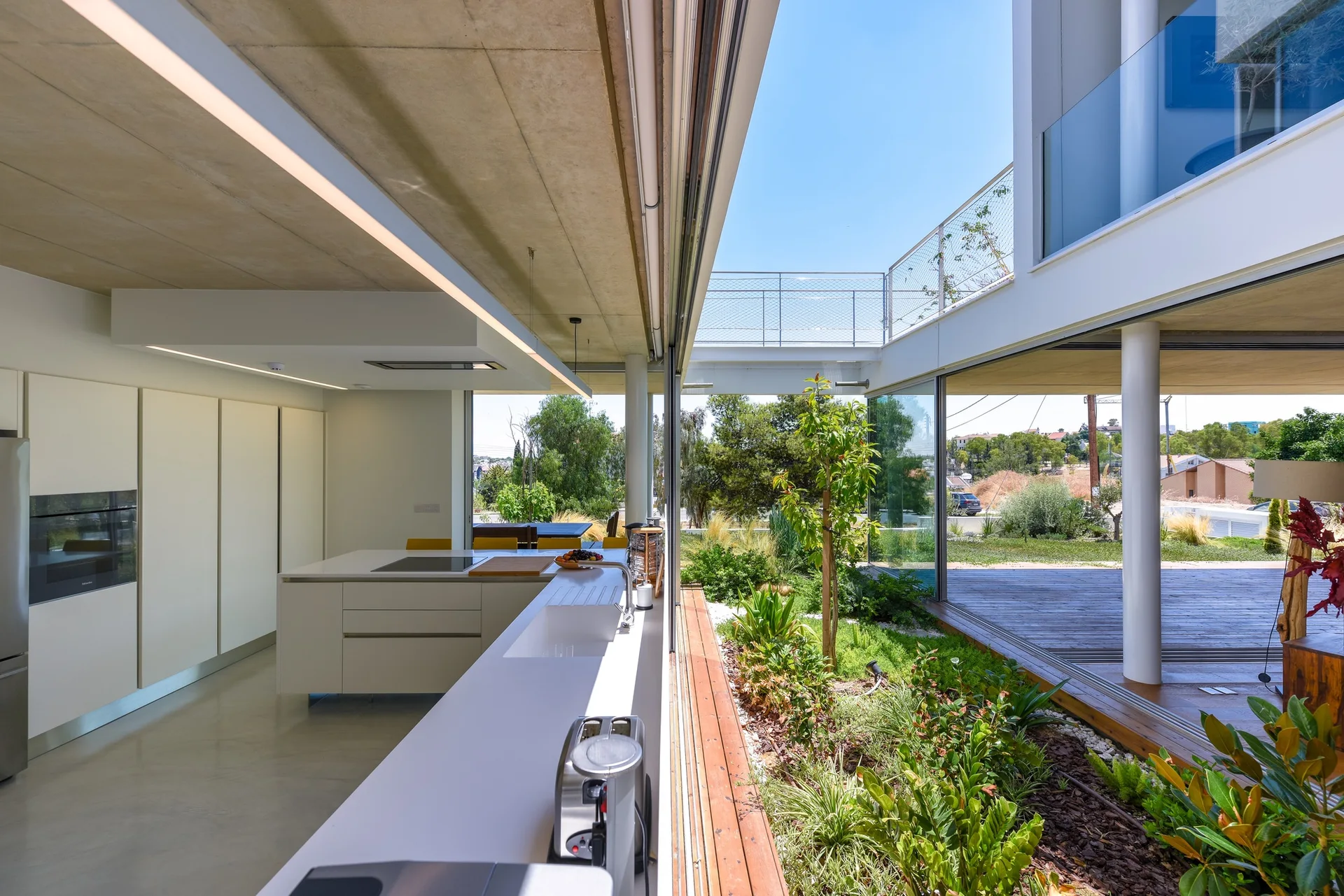 A minimalist white kitchen features flat-panel cabinets, concrete ceilings, and wide sliding glass doors that open to a sunlit central courtyard.