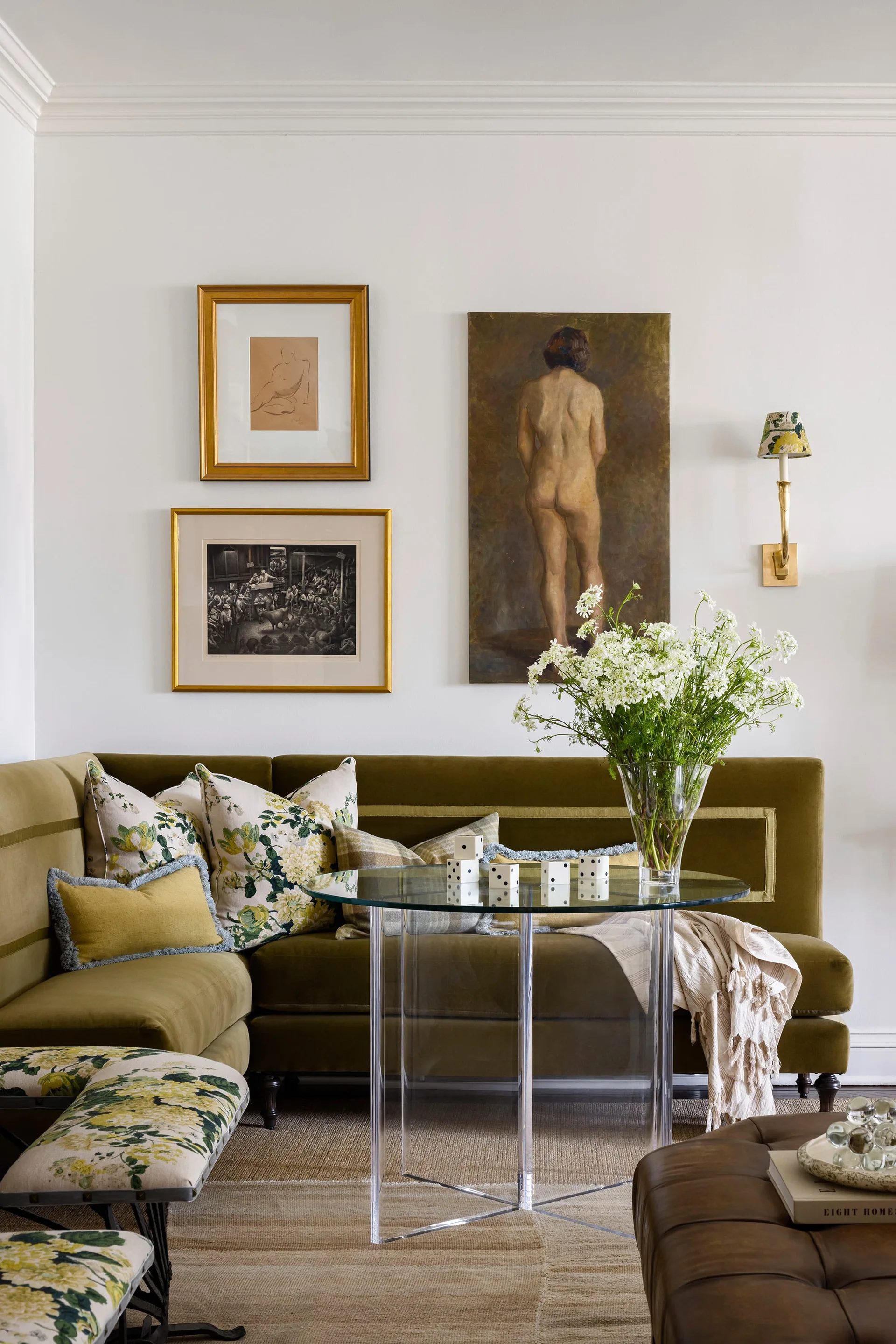 An olive green corner banquette features floral throw pillows behind a round glass table with a clear acrylic base and framed wall art.