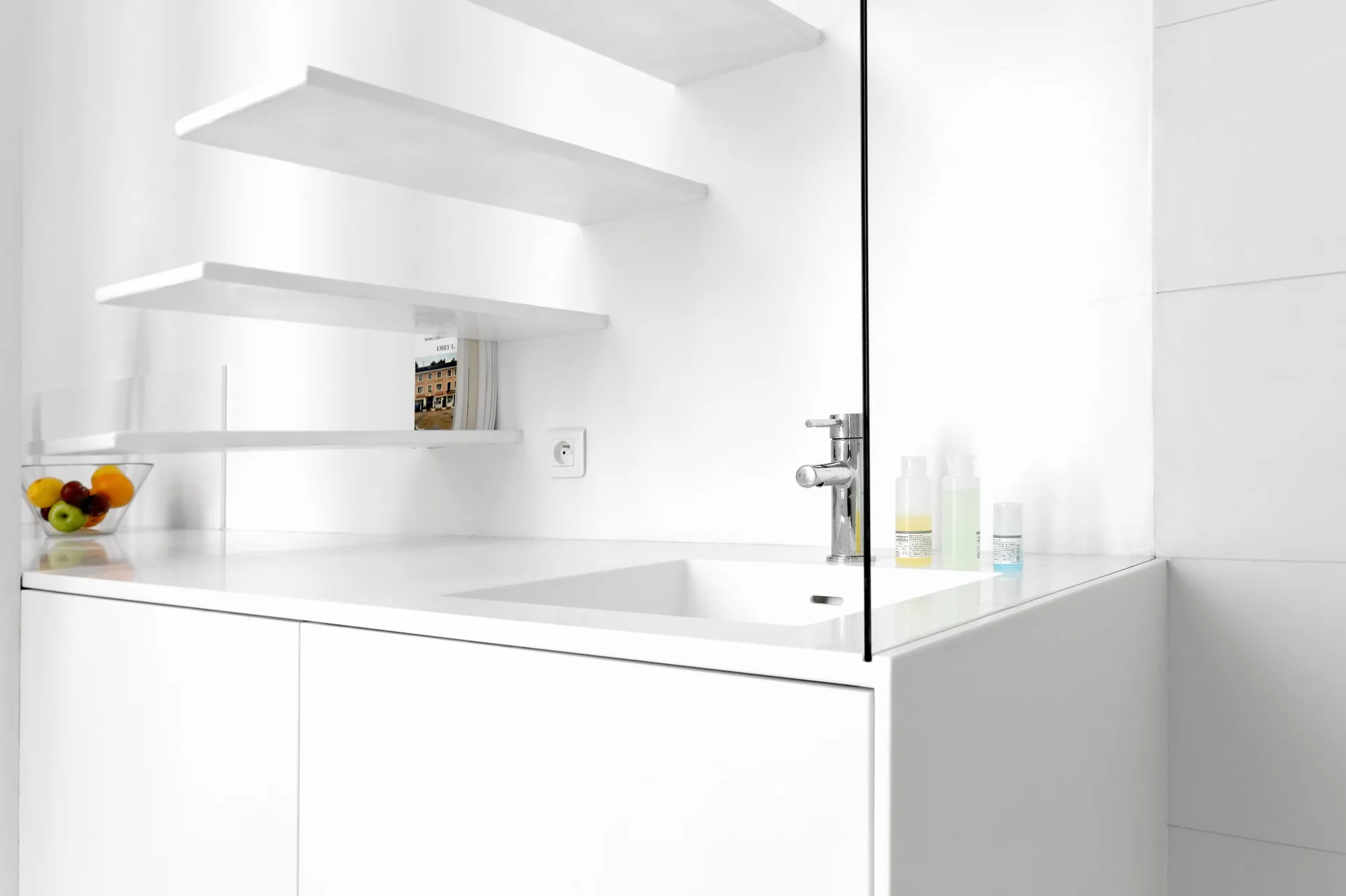 A minimalist white bathroom vanity features an integrated sink with a chrome faucet, a glass splash guard, and tiered white floating shelves on the wall.