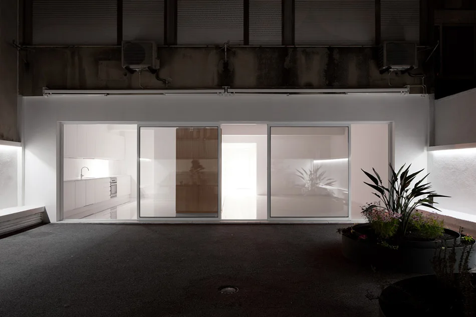 A dark outdoor concrete patio at night features large glass sliding doors looking into a bright, minimalist white kitchen.