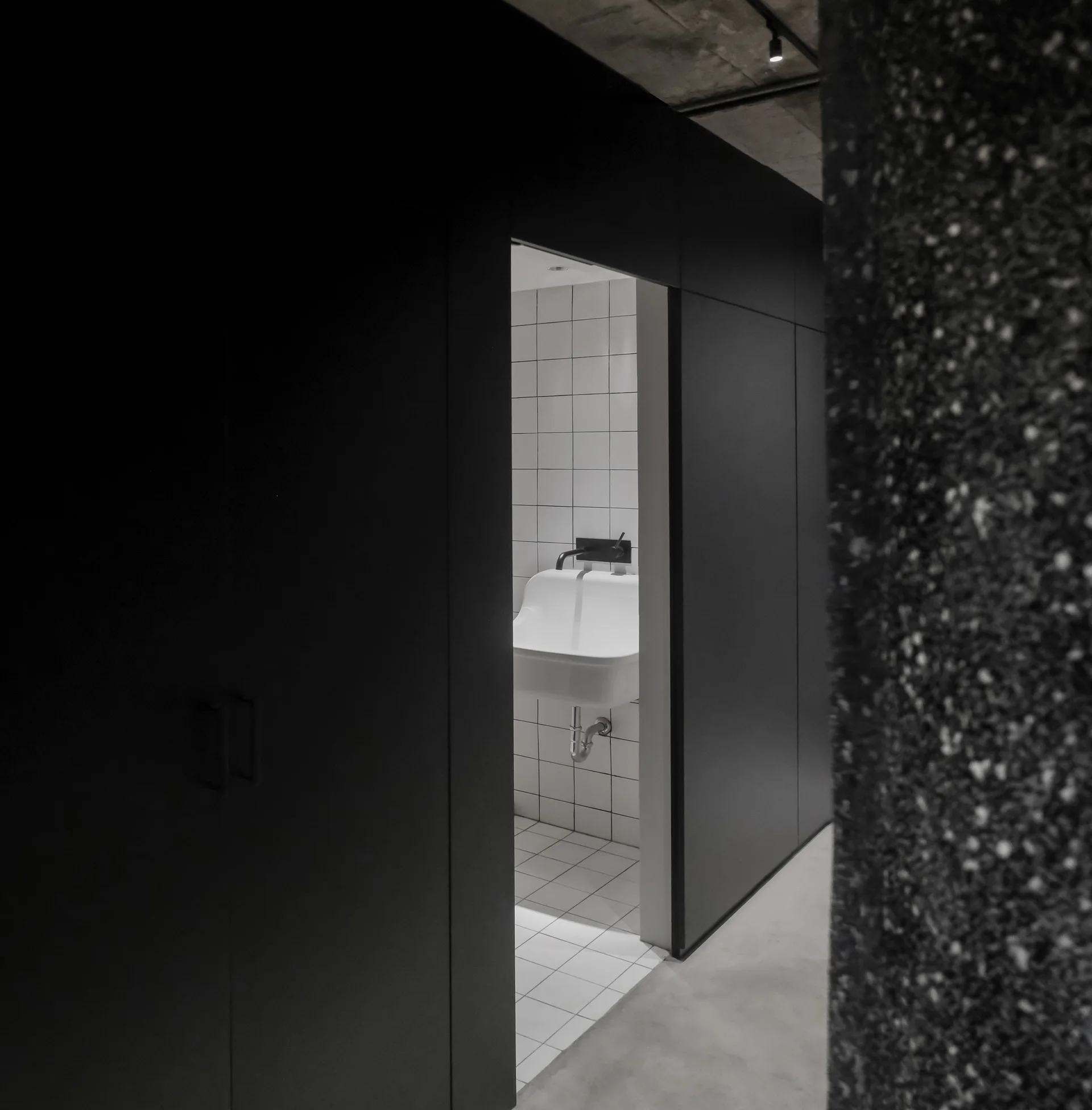 A view through a dark partition wall opening into a bathroom featuring white square grid tiles and a wall-mounted sink.