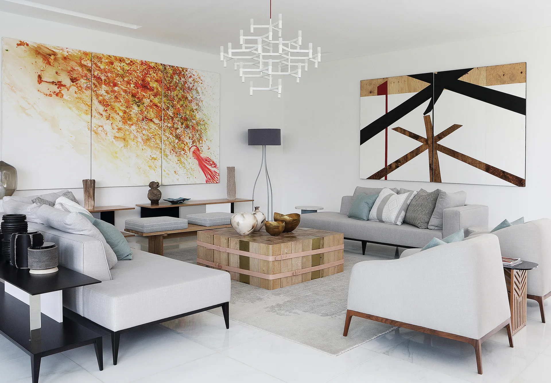 A light gray living room features a blocky wooden coffee table, matching upholstered sofas, large abstract wall art, and a white geometric chandelier.