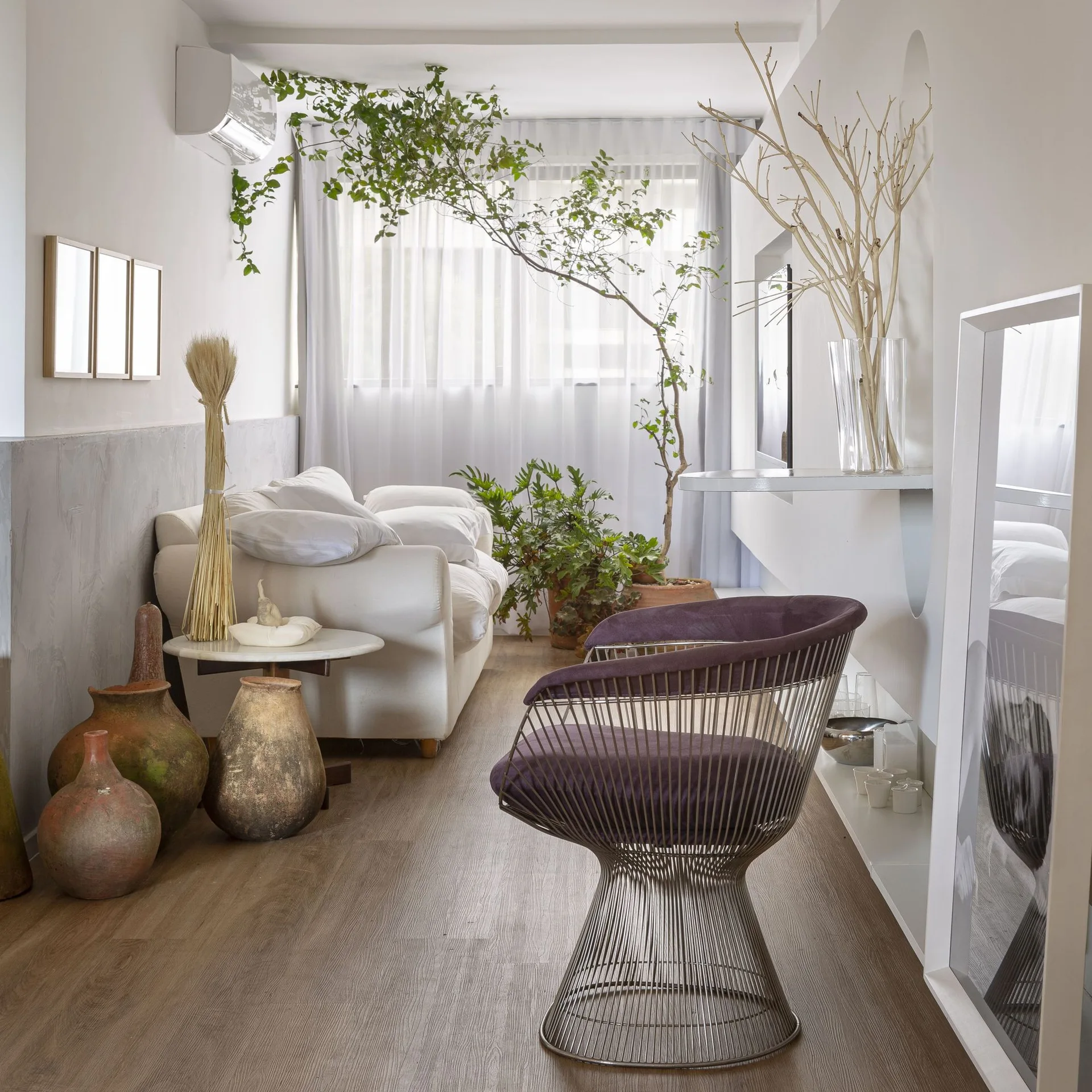 A bright living room features a white sofa, a purple wire-frame armchair, rustic clay vases on the floor, and sheer window curtains.