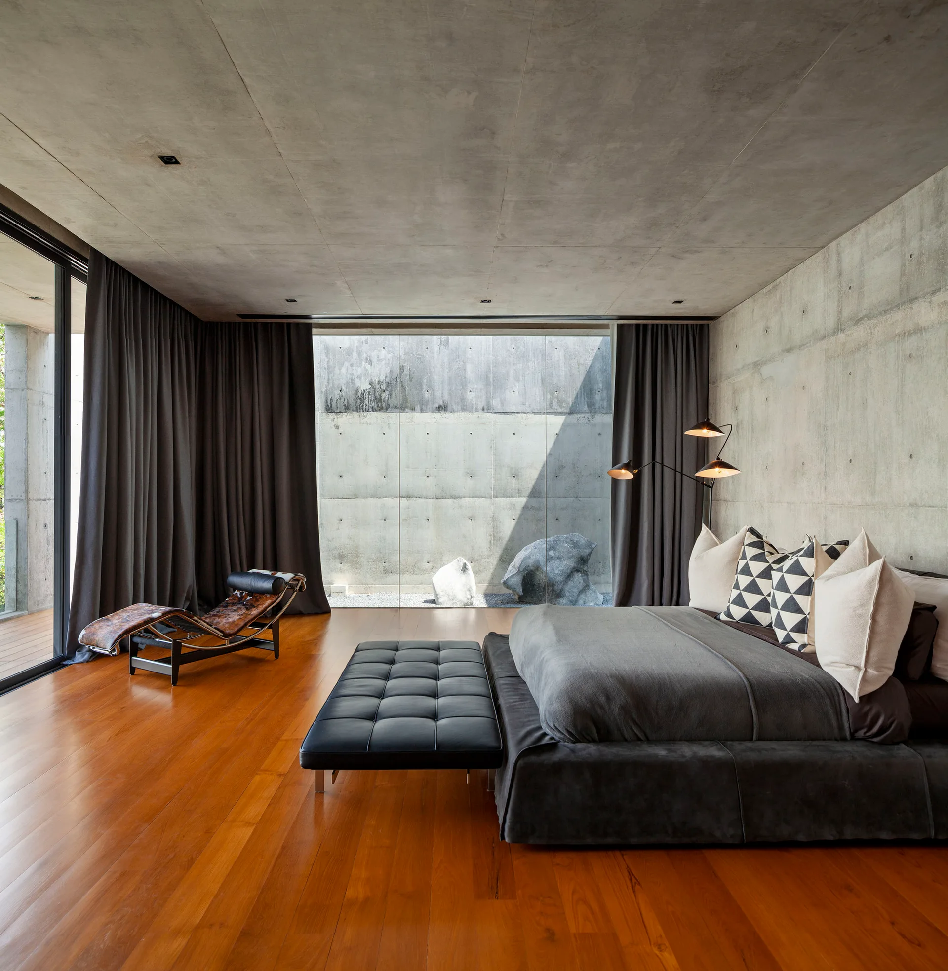 A modern bedroom features raw concrete walls, warm wood flooring, an upholstered bed with patterned pillows, and a black tufted leather bench.