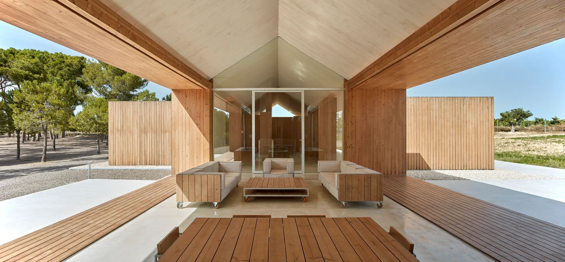 A covered outdoor patio featuring modular wooden sofas, a gabled wood-paneled ceiling, a concrete floor, and a central glass door leading indoors.