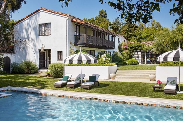 A backyard swimming pool in front of a white stucco house with a dark wood balcony, featuring lounge chairs under black and white striped umbrellas.