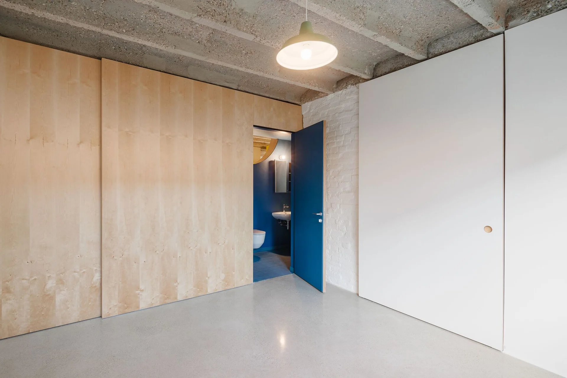 A minimalist hallway featuring plywood wall paneling, a concrete floor and ceiling, and an open blue door revealing a matching blue bathroom.