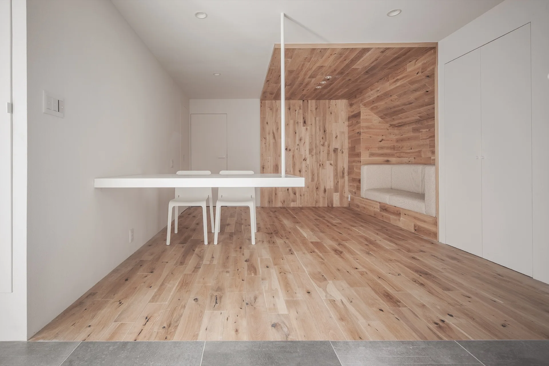 A minimalist dining space features a white cantilevered table with two white chairs alongside a built-in bench set inside a natural wood alcove.