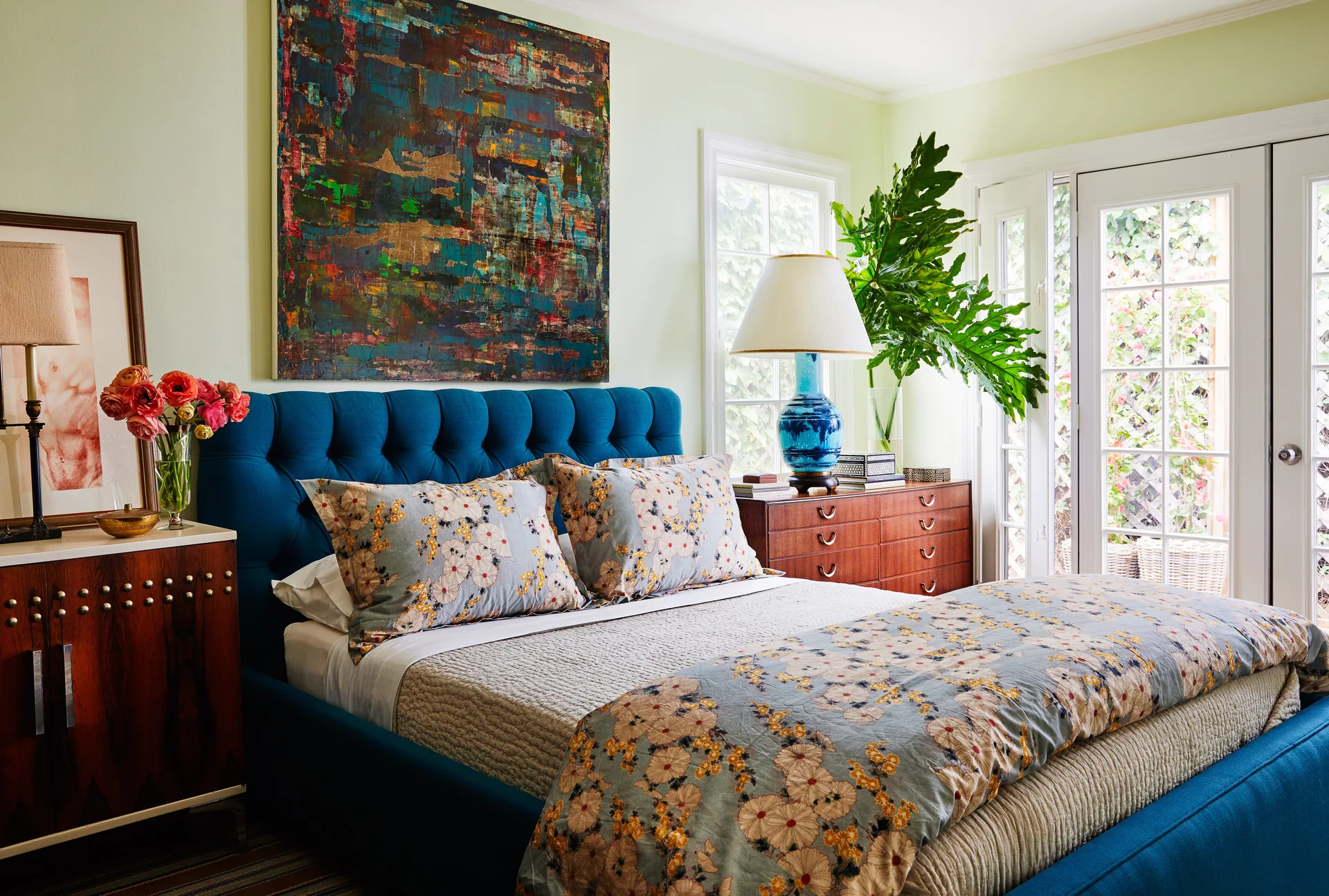 An eclectic bedroom features a bright blue tufted headboard, floral patterned bedding, a large abstract painting, and mid-century wooden dressers.