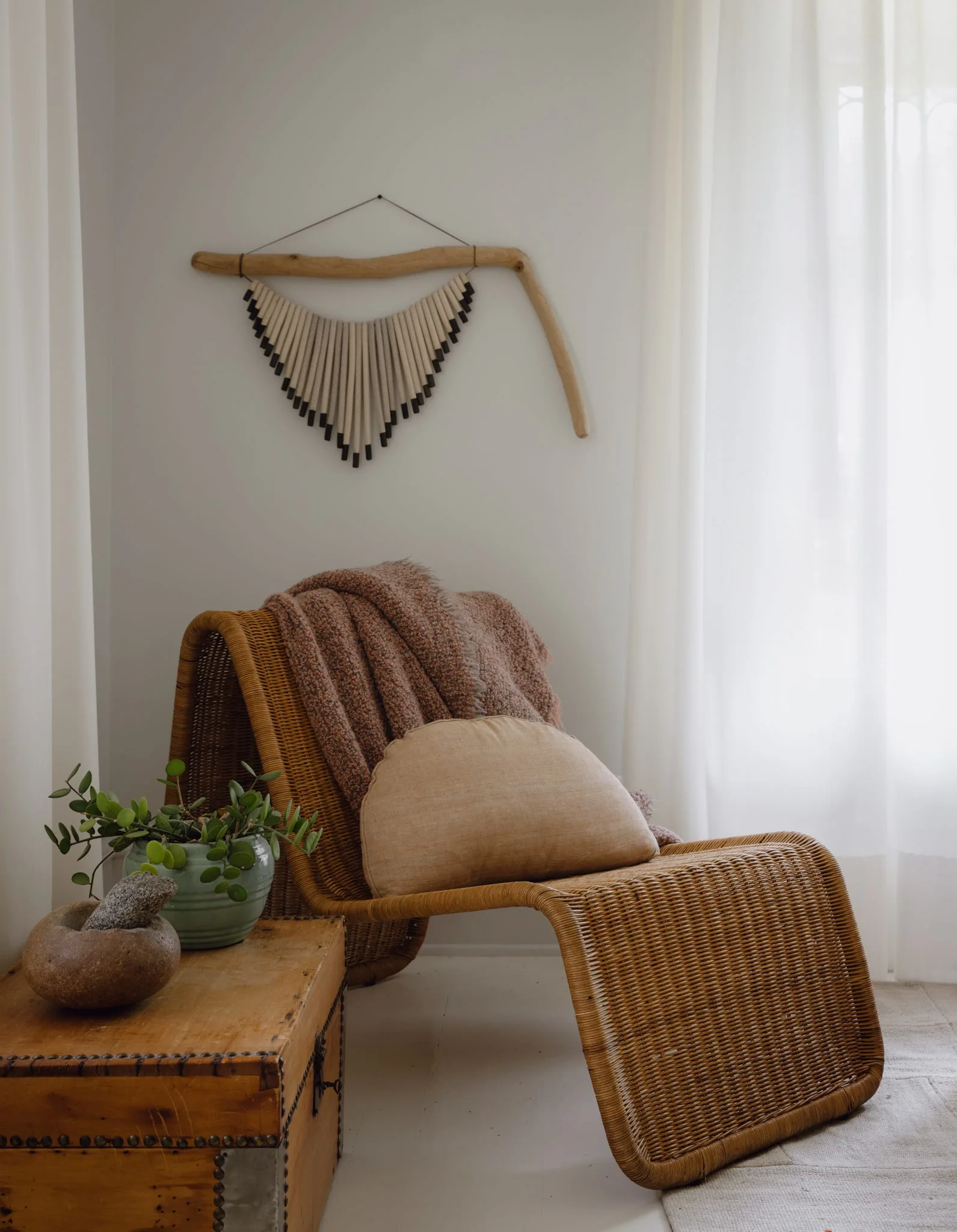 A curved wicker lounge chair with a throw blanket sits in a sunlit corner next to a wooden chest and driftwood wall art.