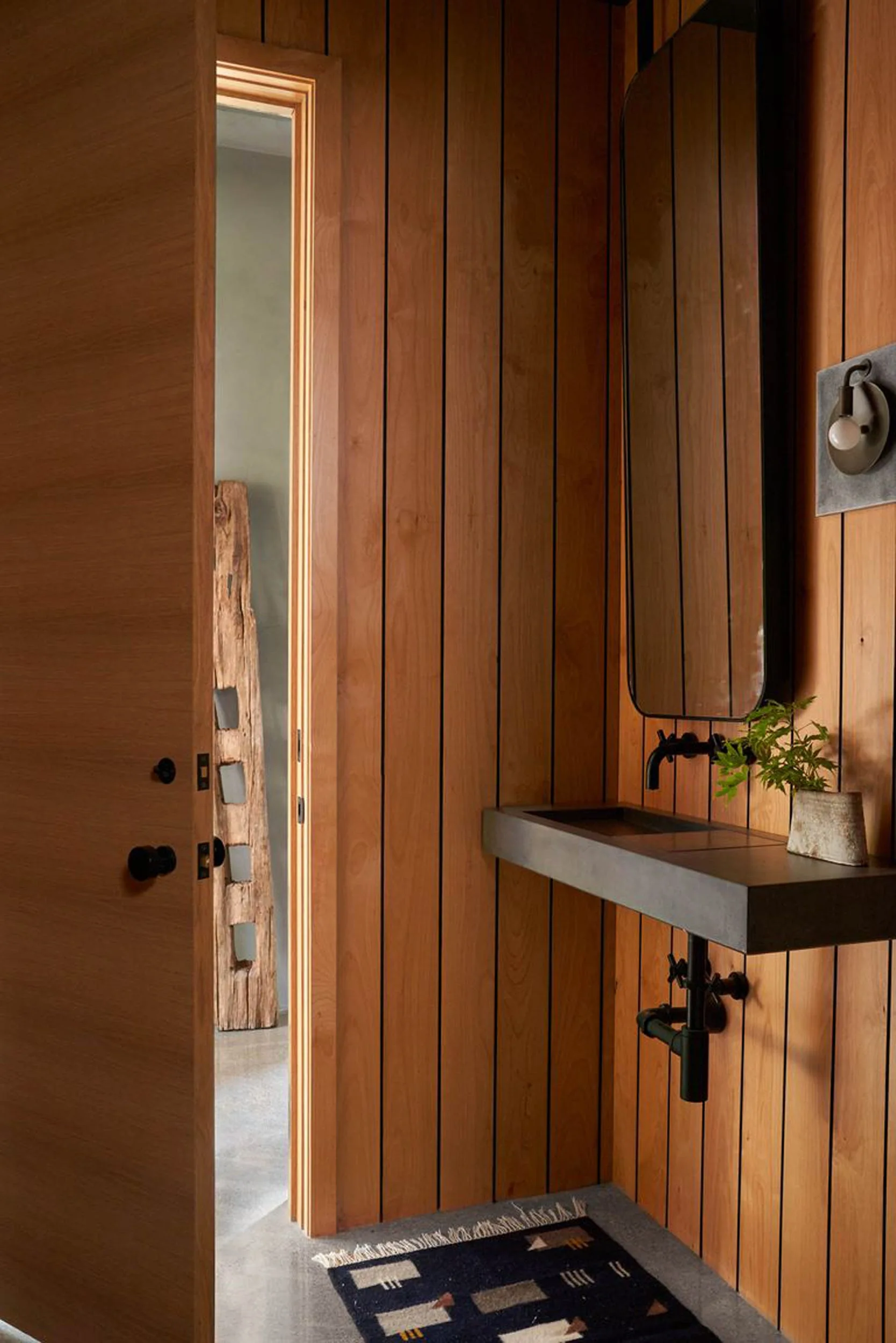 A modern powder room features vertical wood plank walls, a floating concrete sink with a black faucet, and a tall thin mirror.