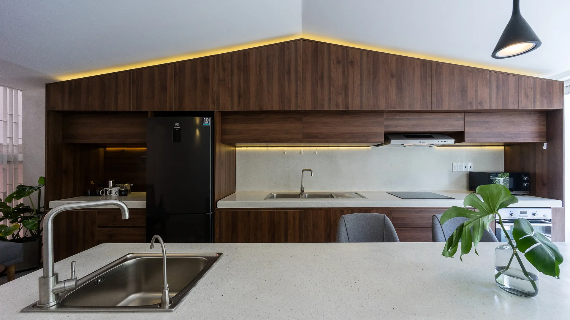 A modern kitchen featuring wood-grained cabinets cut to match a sloped ceiling, illuminated by LED lights above a white speckled countertop.