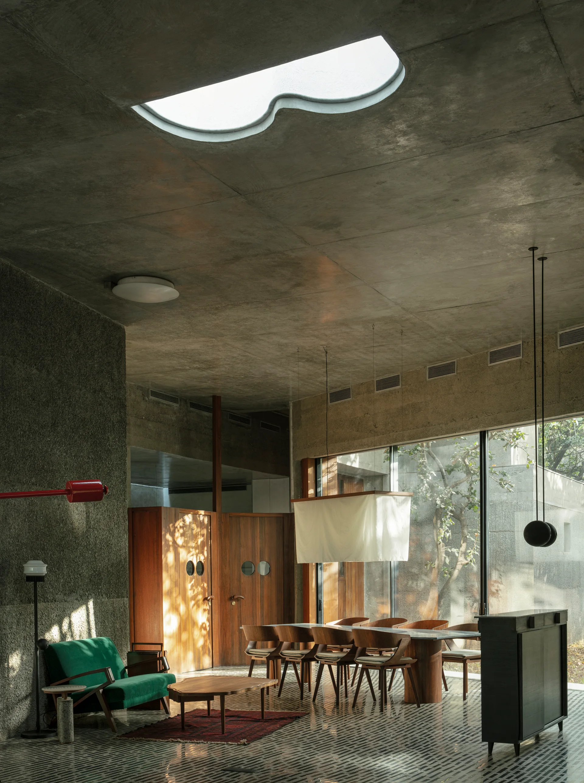 A dining and living space features a concrete ceiling with a curved skylight, a wooden dining set, and a green sofa near large glass windows.