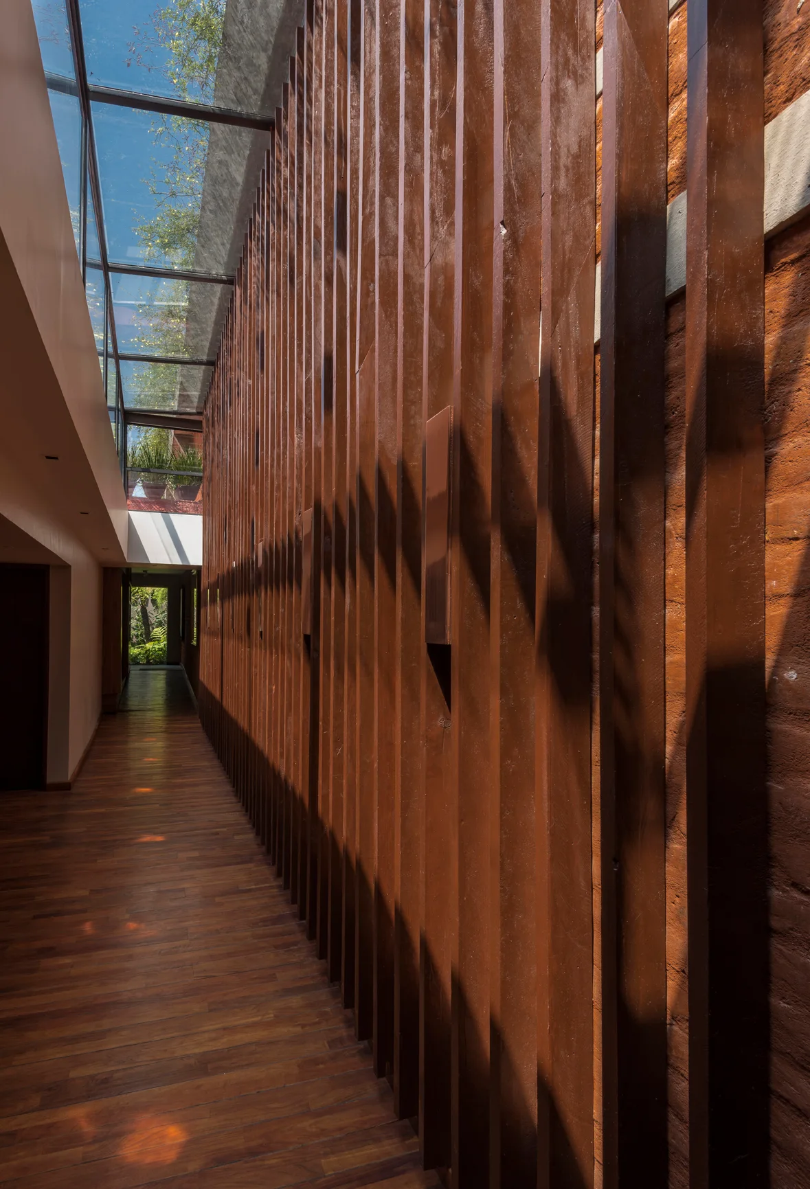 A long indoor hallway features dark vertical wood slats along one wall under a glass skylight roof and wood flooring.