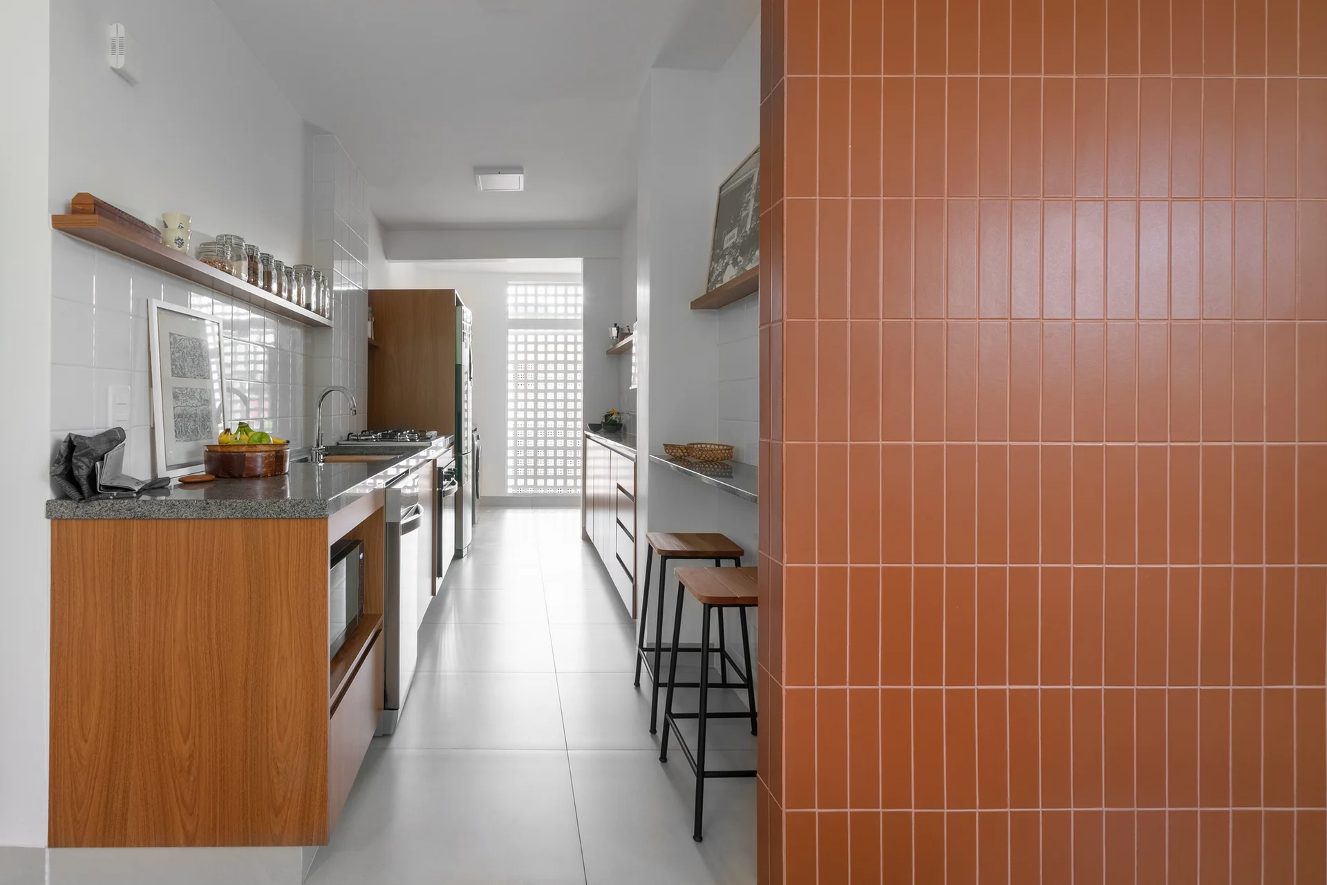 A modern galley kitchen featuring wood cabinetry, grey countertops, bar stools, and a prominent terracotta tiled wall partition in the foreground.