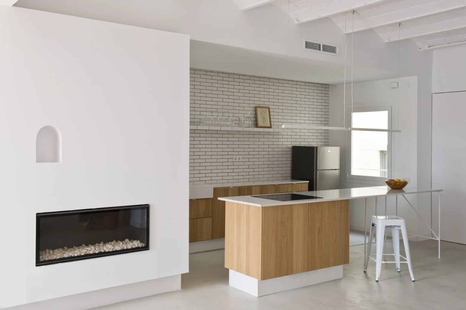 A minimalist kitchen features a wood-toned island, a white tiled backsplash, a metal stool, and a modern fireplace built into a white partition wall.