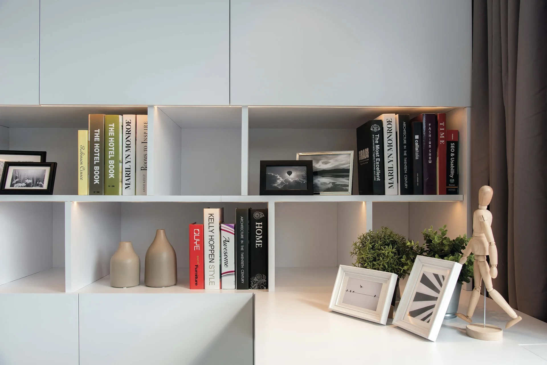 A white built-in shelving unit features books, small framed pictures, two ceramic vases, potted green plants, and a wooden mannequin figure.