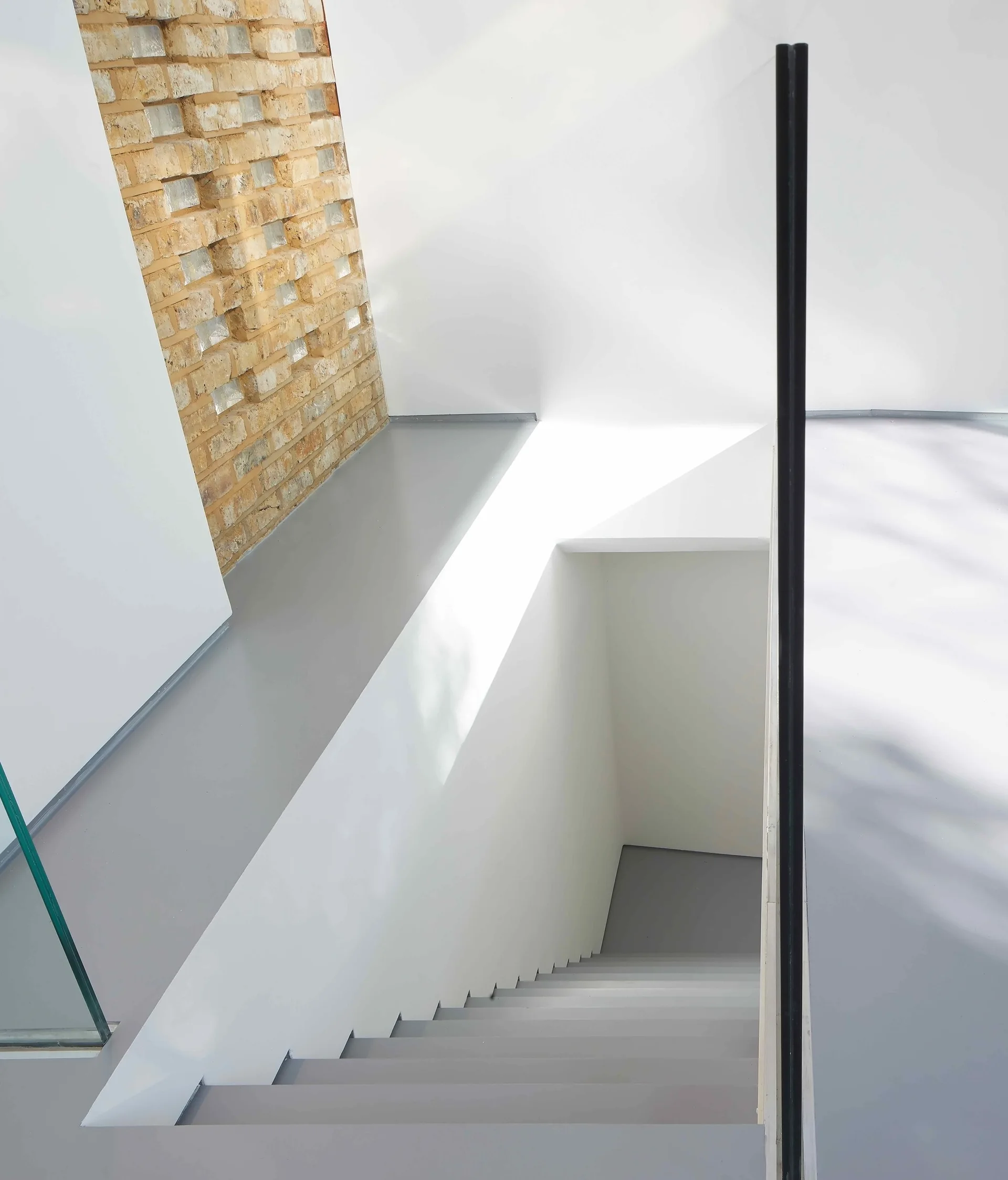 A minimalist staircase with matte grey steps descending between white walls and an exposed brick accent wall under bright natural light.
