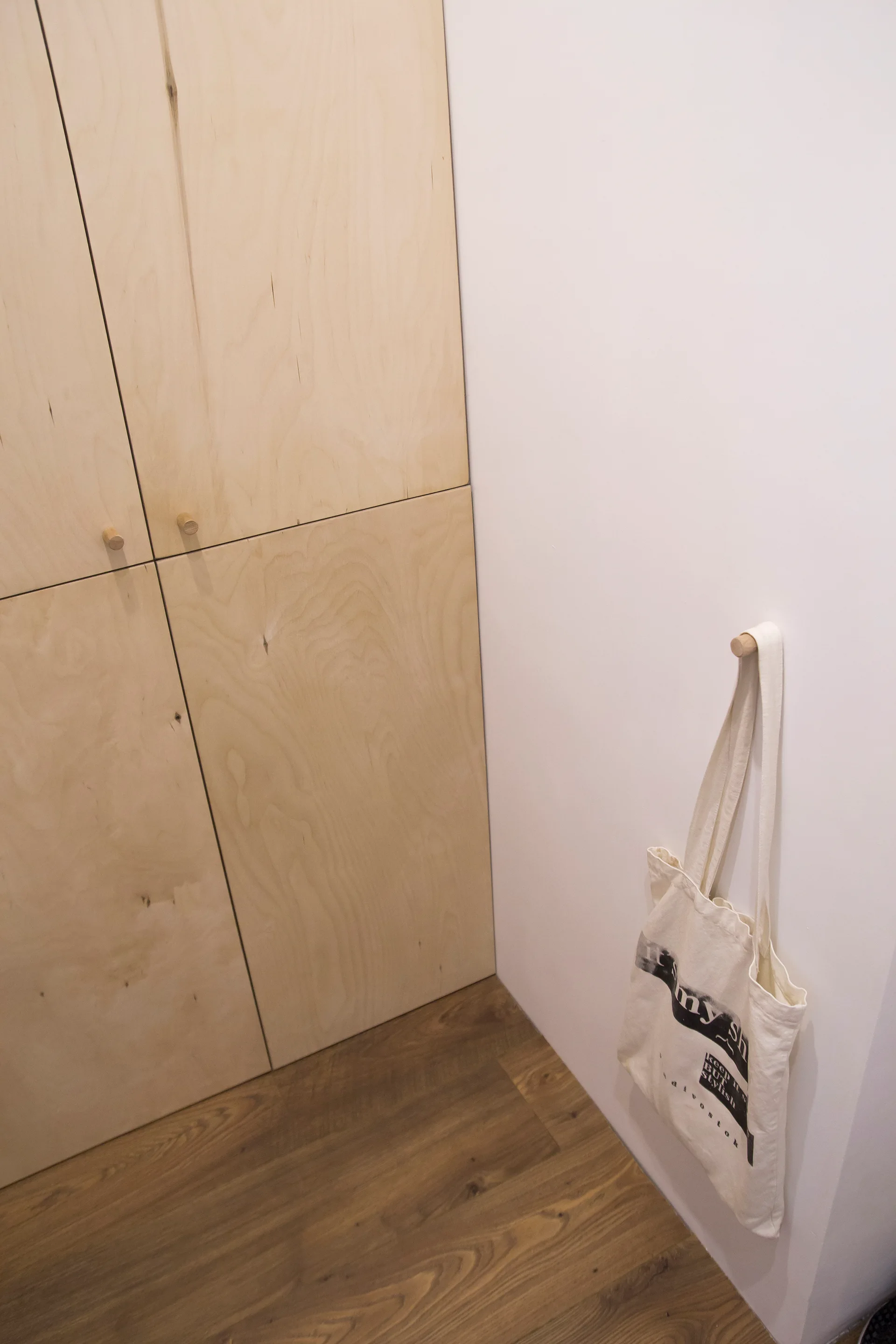 Built-in plywood closet doors with round wooden knobs sit adjacent to a smooth white wall holding a hanging canvas tote bag.