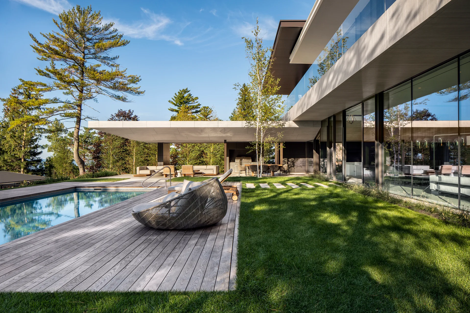 A rectangular swimming pool is flanked by a weathered wooden deck, a green lawn, and a modern home with a glass facade.