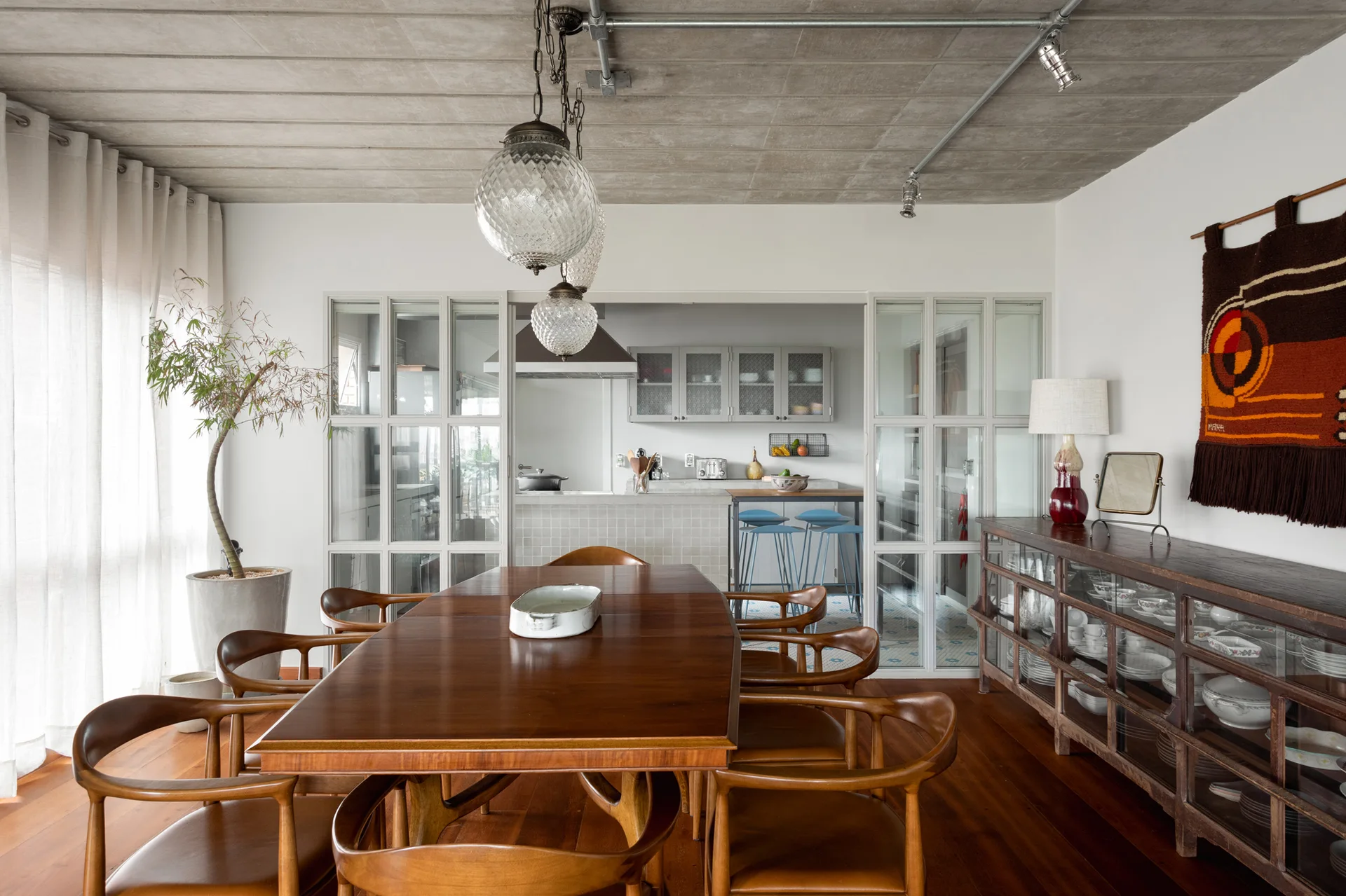 A wooden dining table and chairs sit beneath an exposed concrete ceiling, with glass-paned sliding doors opening to an adjacent kitchen.