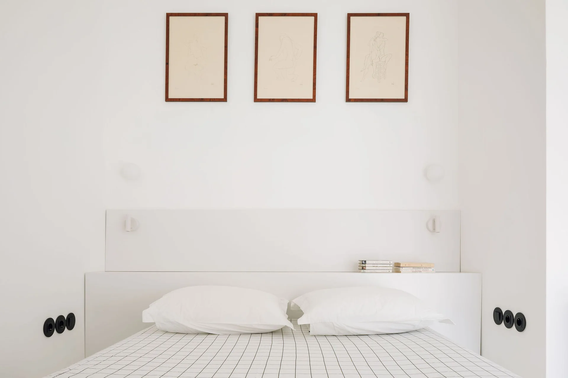 A minimalist white bedroom features a bed with black and white grid bedding, an integrated white ledge headboard, and three framed sketches.