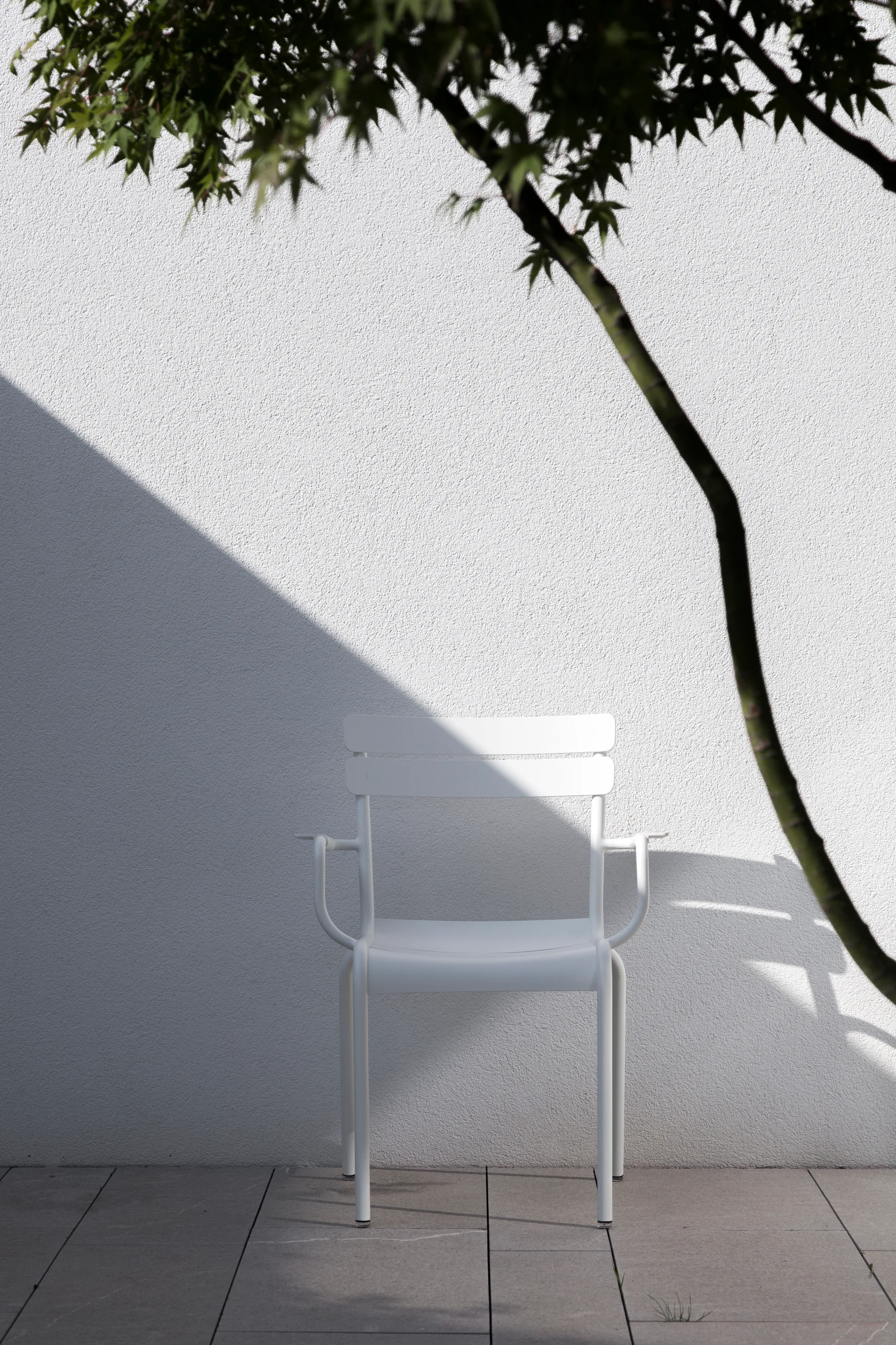 A white metal outdoor armchair stands on a grey stone patio against a textured white wall cast with sharp diagonal shadows.