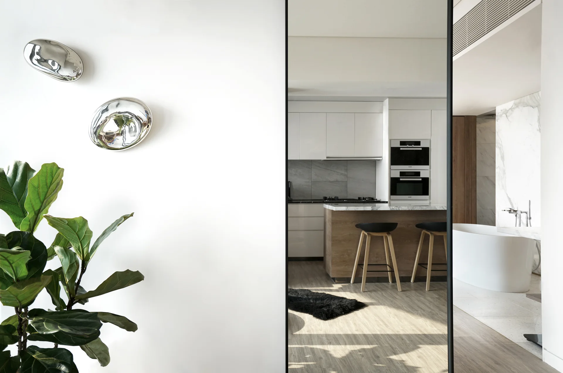 A modern interior view through a black framed opening showing a kitchen with a wooden island and a bathroom with a white bathtub.