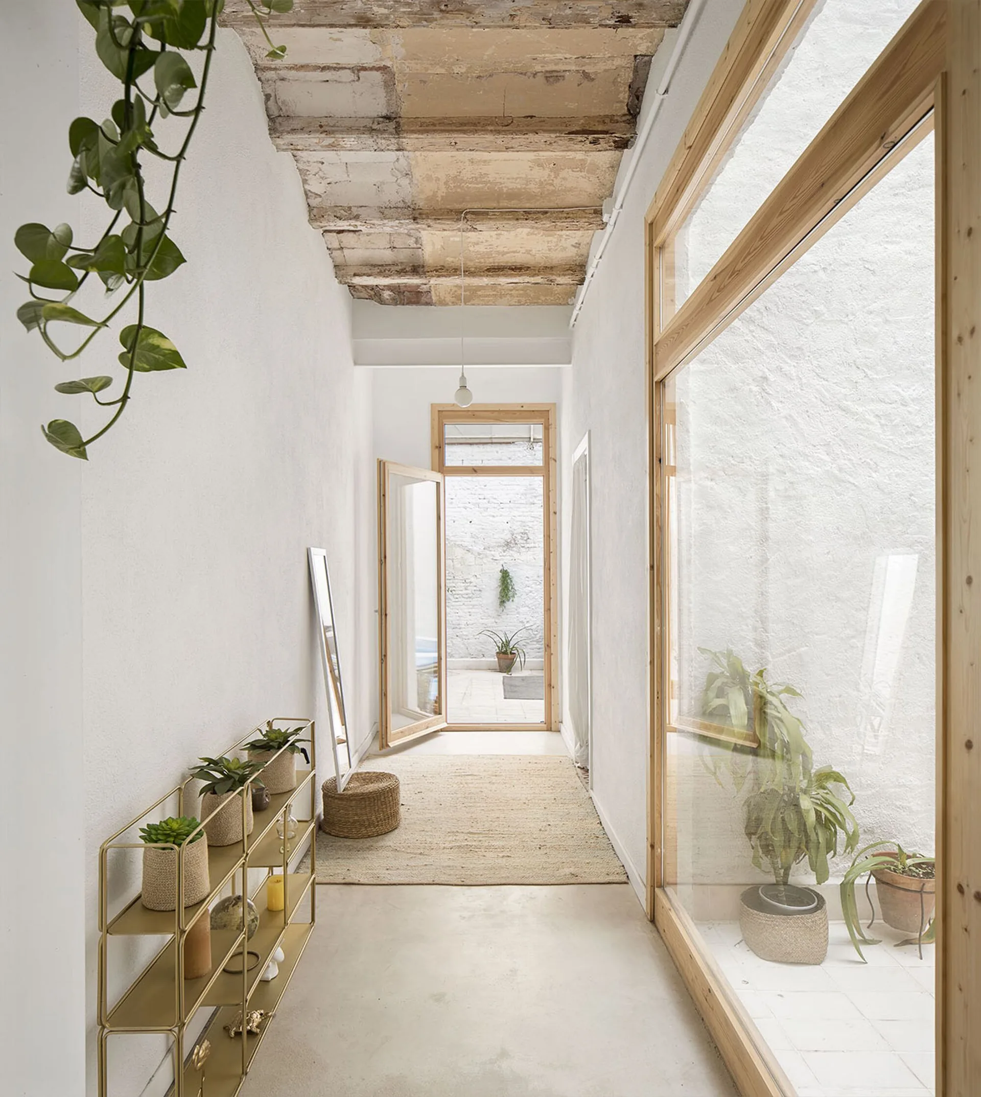A narrow white hallway featuring a raw exposed ceiling, a gold metal shelf with plants, and large wood-framed glass doors looking into a courtyard.