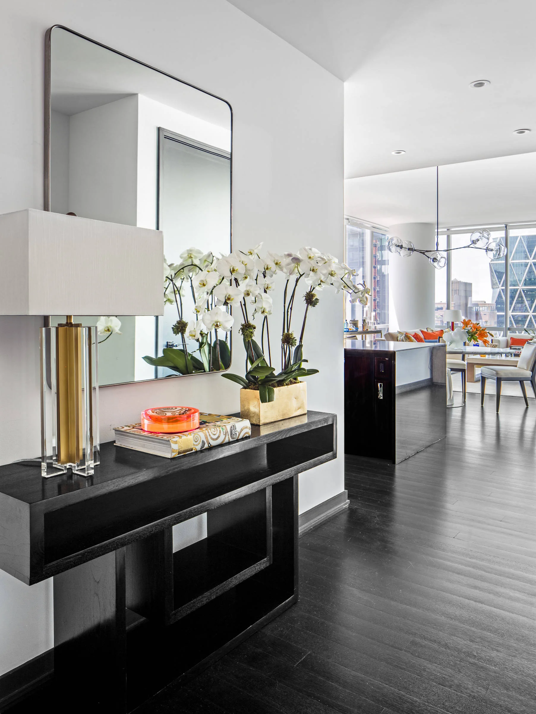 A modern entryway featuring a black wood console table with a large mirror, a gold-base lamp, and white orchids on dark wood floors.