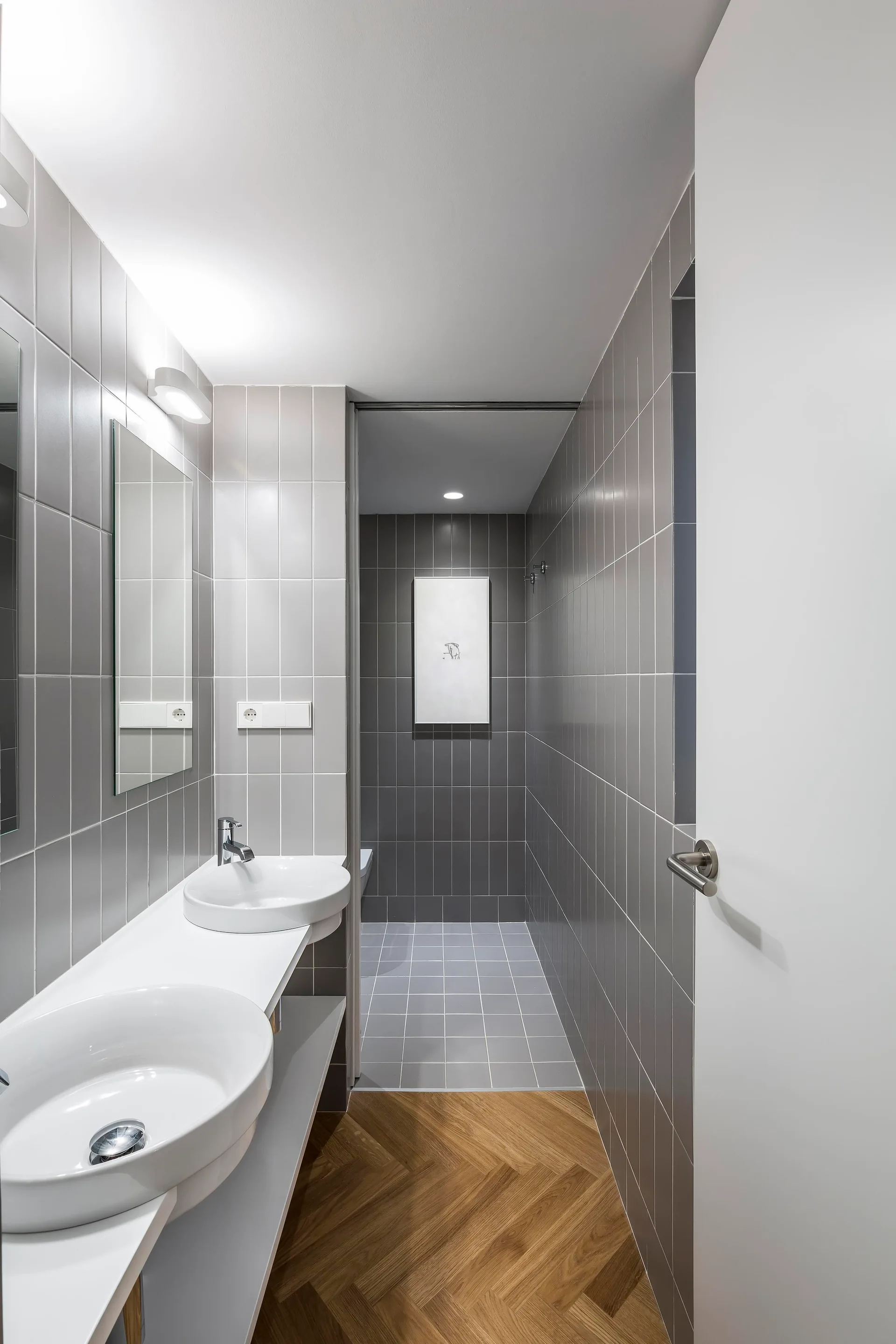 A narrow modern bathroom features a white double vanity, grey vertical tiled walls, and herringbone pattern wood flooring leading to a walk-in shower.