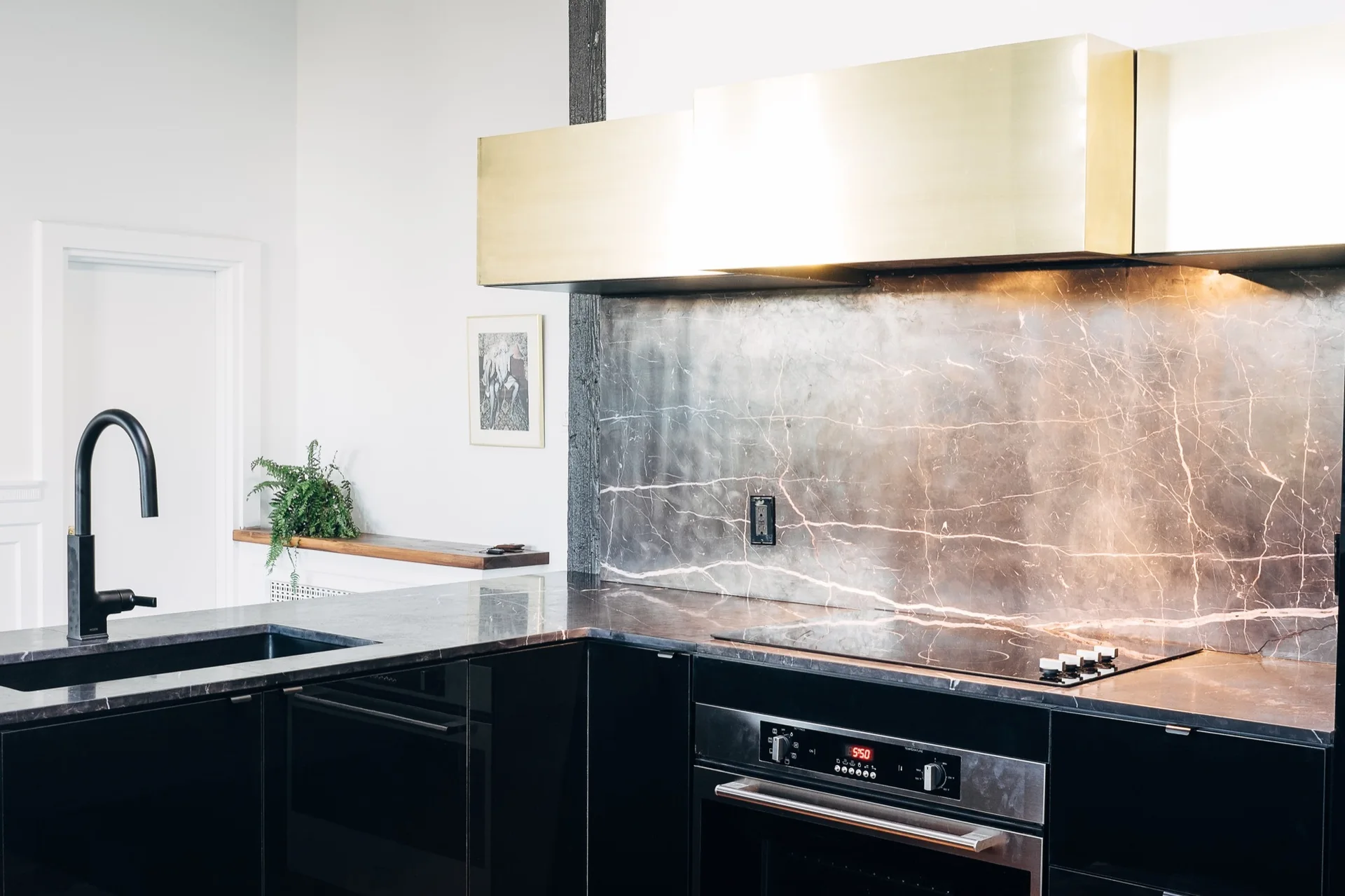 A modern kitchen featuring black glossy lower cabinets, a dark veined marble backsplash and countertop, and a large brass range hood.