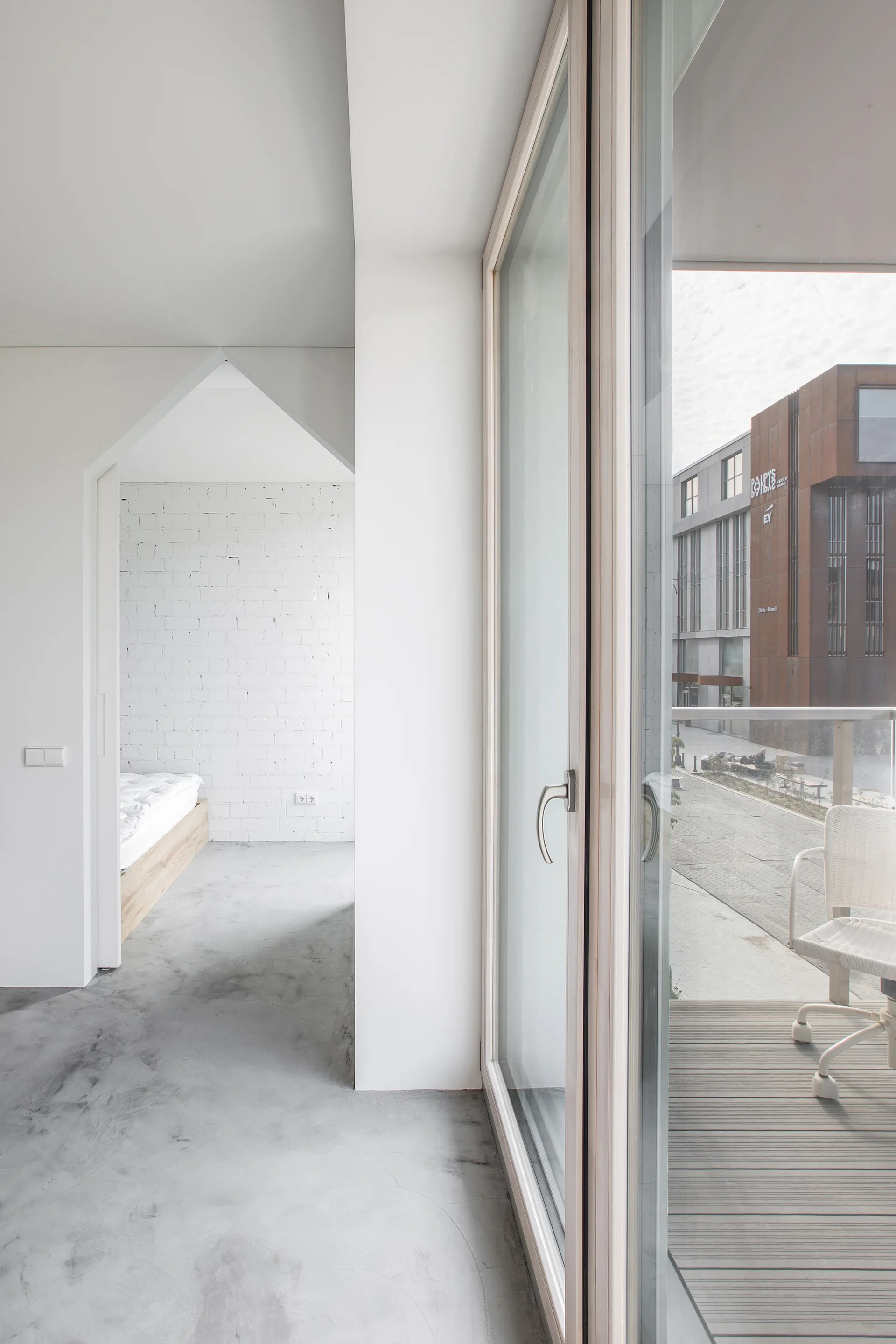 A minimalist interior with concrete flooring features a sloped doorway leading to a bedroom and a large glass door opening to a balcony.
