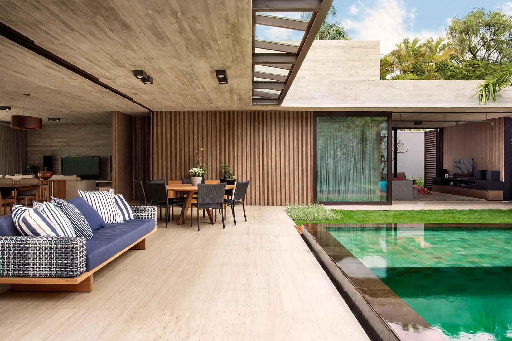 A modern covered patio features a raw concrete ceiling, a wood-slatted accent wall, a blue outdoor sofa, and a turquoise swimming pool.