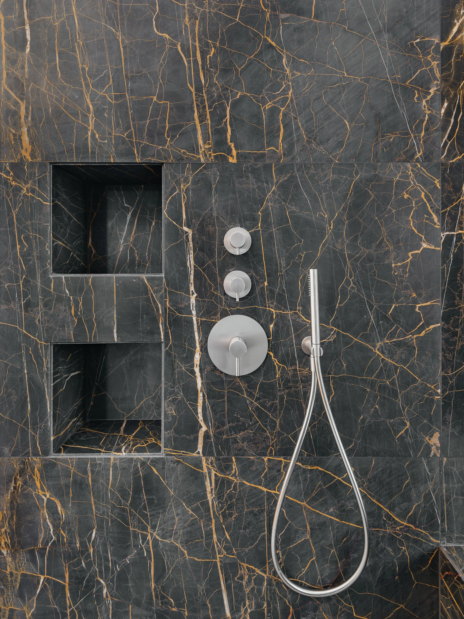 A close-up of a shower wall lined with gold-veined black marble tile, featuring brushed metal controls and two stacked recessed storage niches.