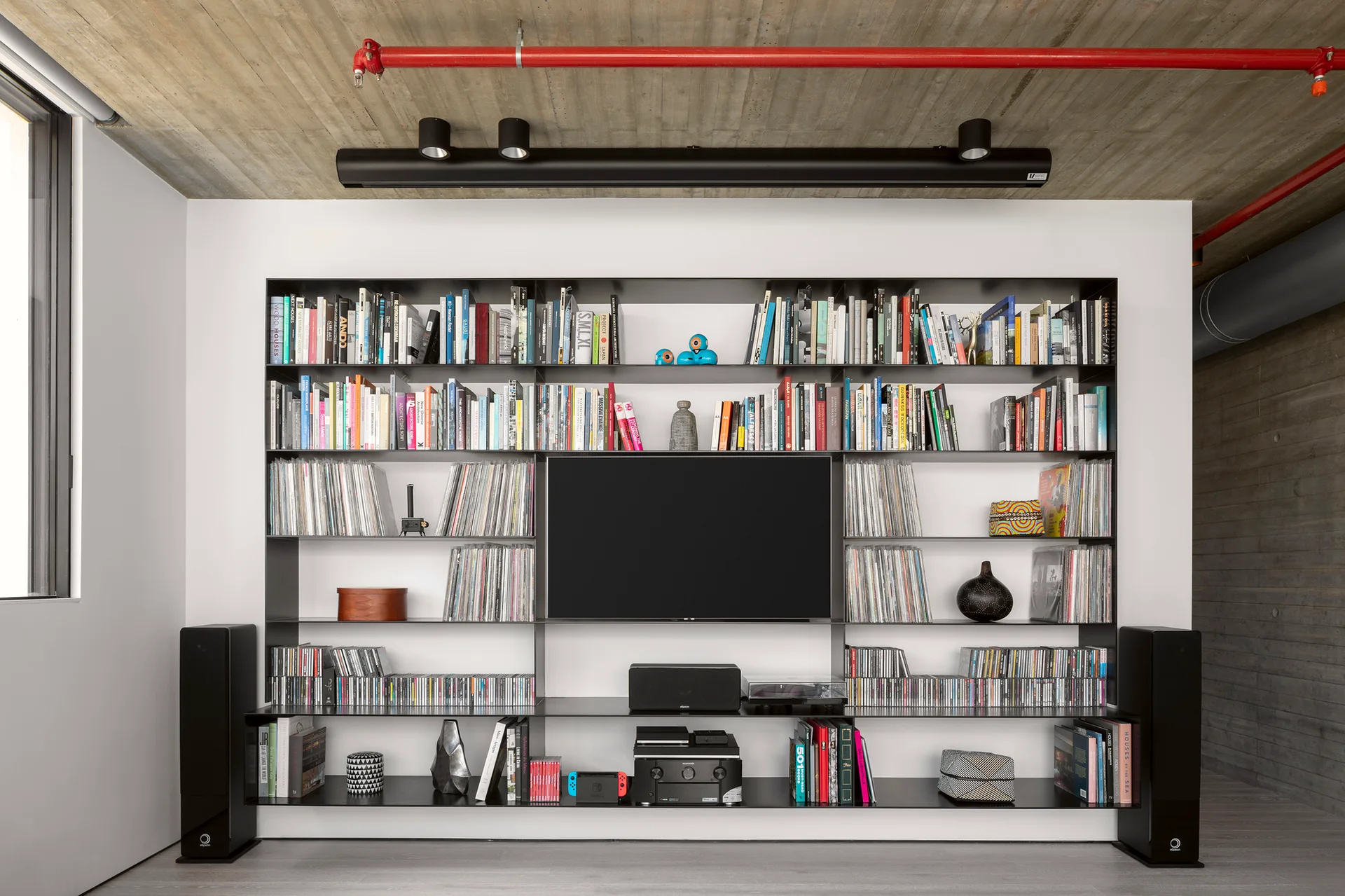 A custom black metal shelving unit holding books and vinyl records surrounds a TV below a raw concrete ceiling.